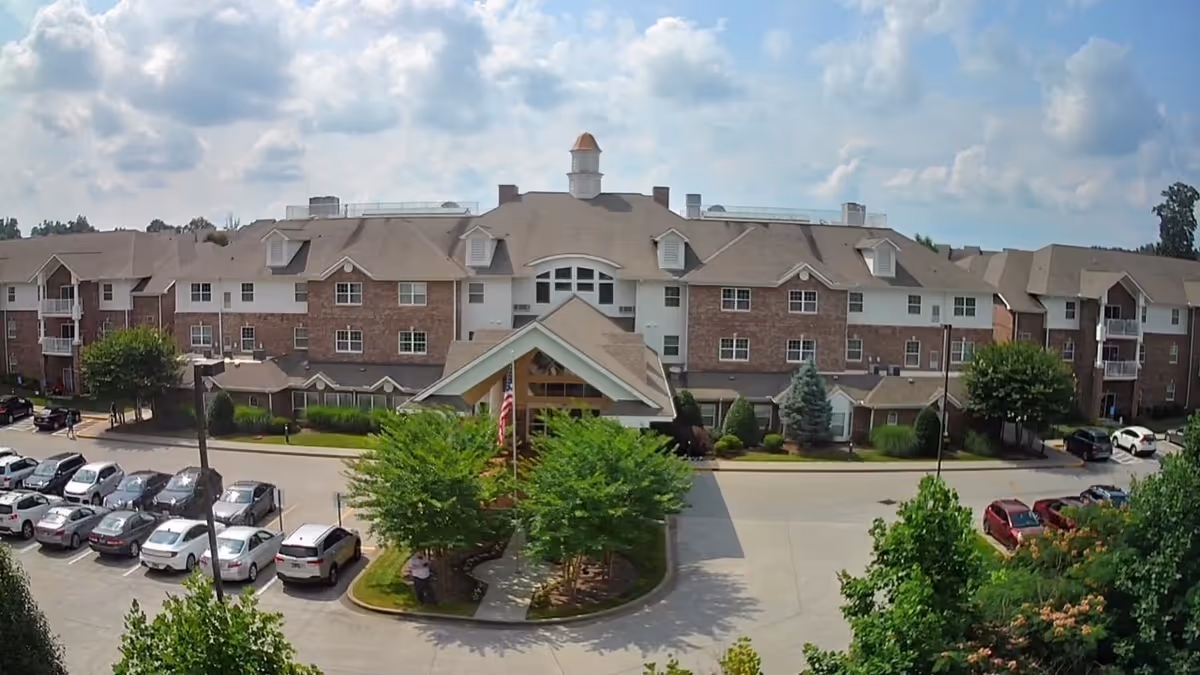 Front exterior view of Sherrill Hills Retirement Resort, a large multi-story brick and siding building with a peaked entrance, surrounded by trees and a parking lot filled with cars under a partly cloudy sky.