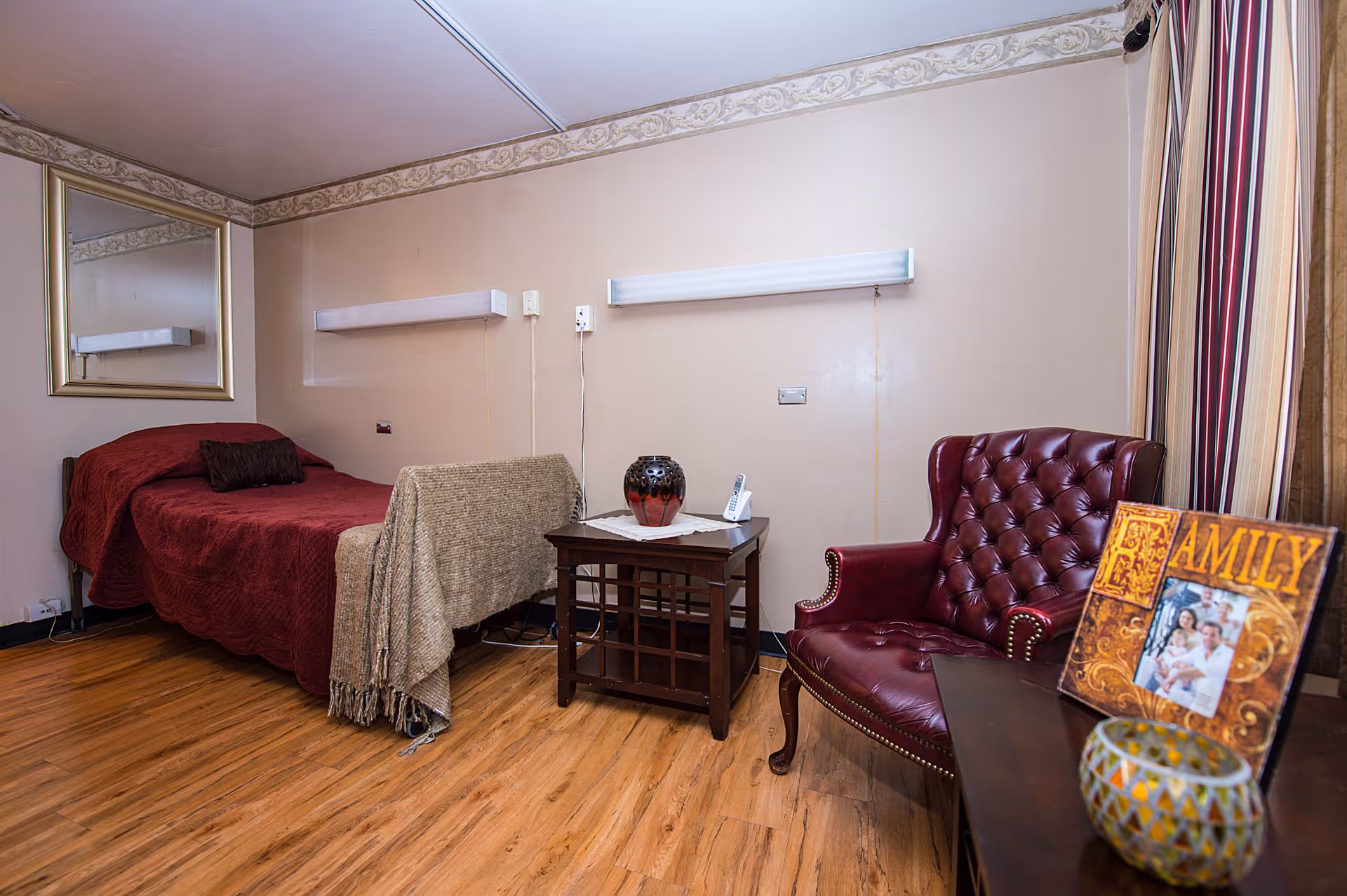 A furnished senior care bedroom with a single bed covered in red bedding, a side table with a vase and phone, and a red leather armchair.