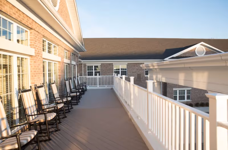 A spacious outdoor balcony area at Liberty Station Health Campus with a row of black rocking chairs featuring striped cushions along the left side. The balcony has white railings and overlooks a brick building under a clear blue sky.
