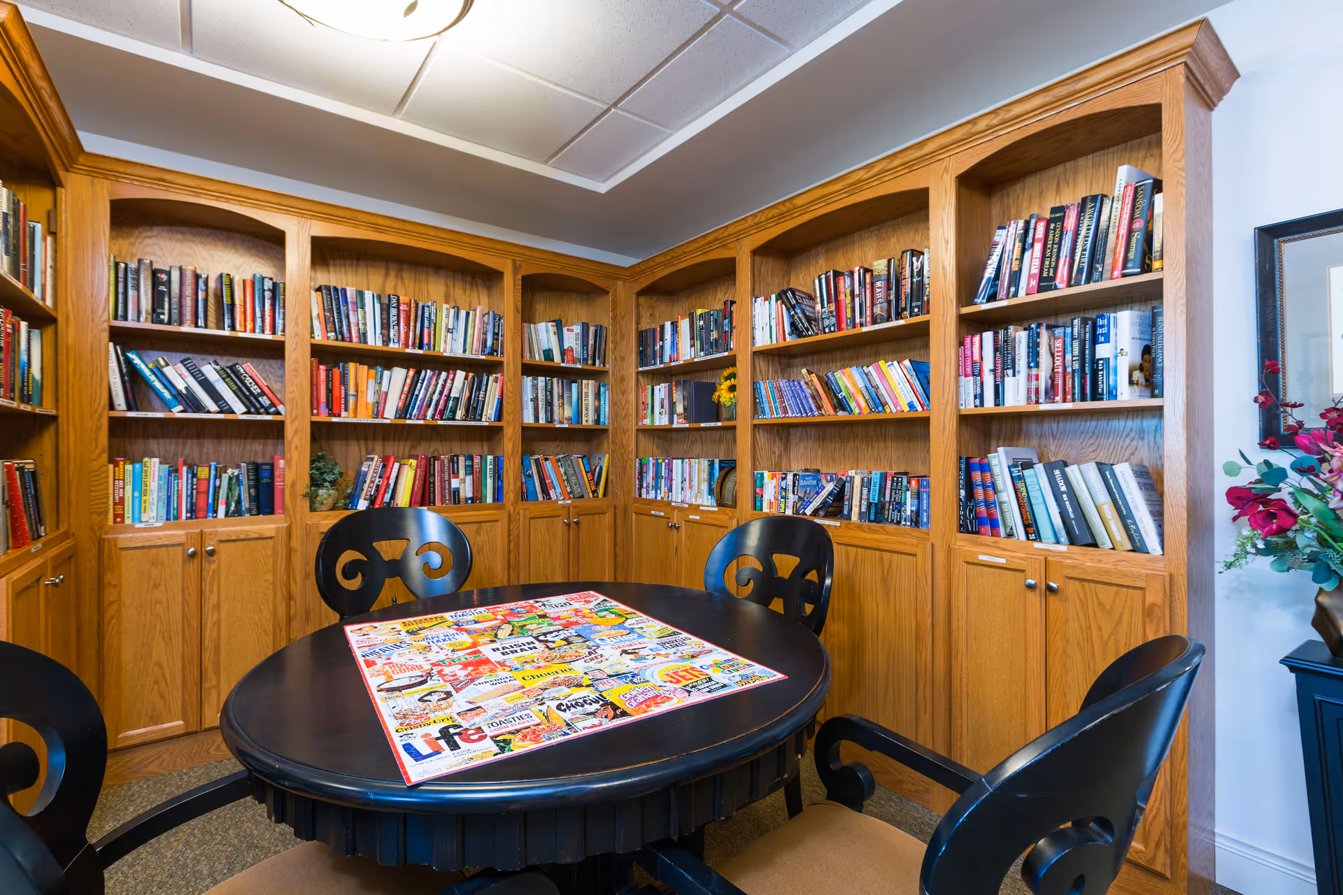 A cozy reading room with wooden bookshelves filled with books lining the walls. In the center, there is a round black table with a colorful puzzle on top and four black chairs with cushioned seats around it. A vase with flowers is visible on a small table to the right.