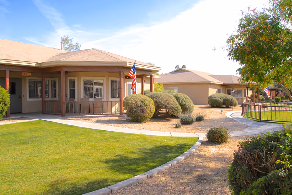 Single-story senior living building with a covered front porch, manicured lawn, walkways, and American flags.