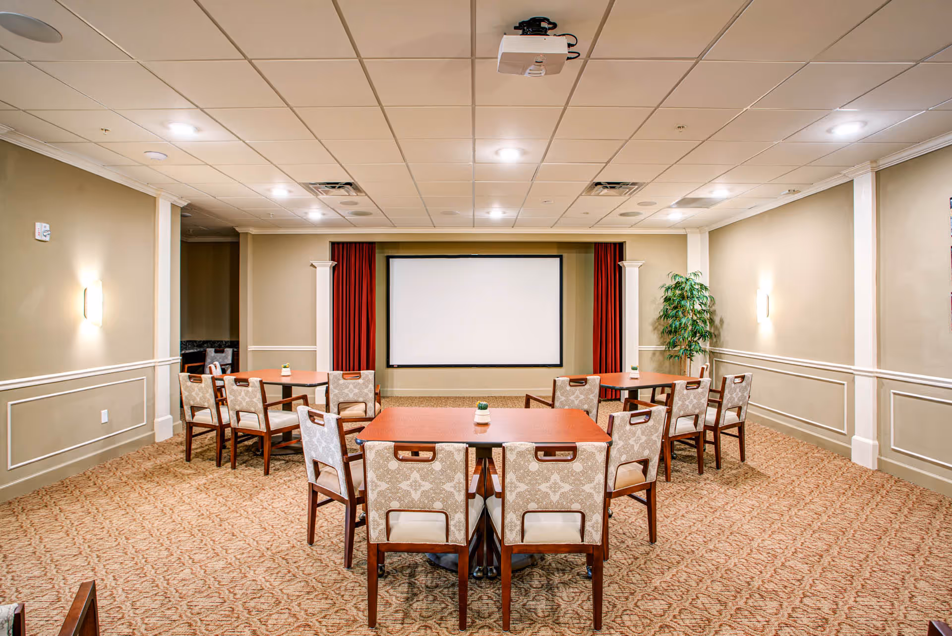 A carpeted multipurpose meeting room with tables and upholstered chairs arranged facing a large projection screen with red curtains.