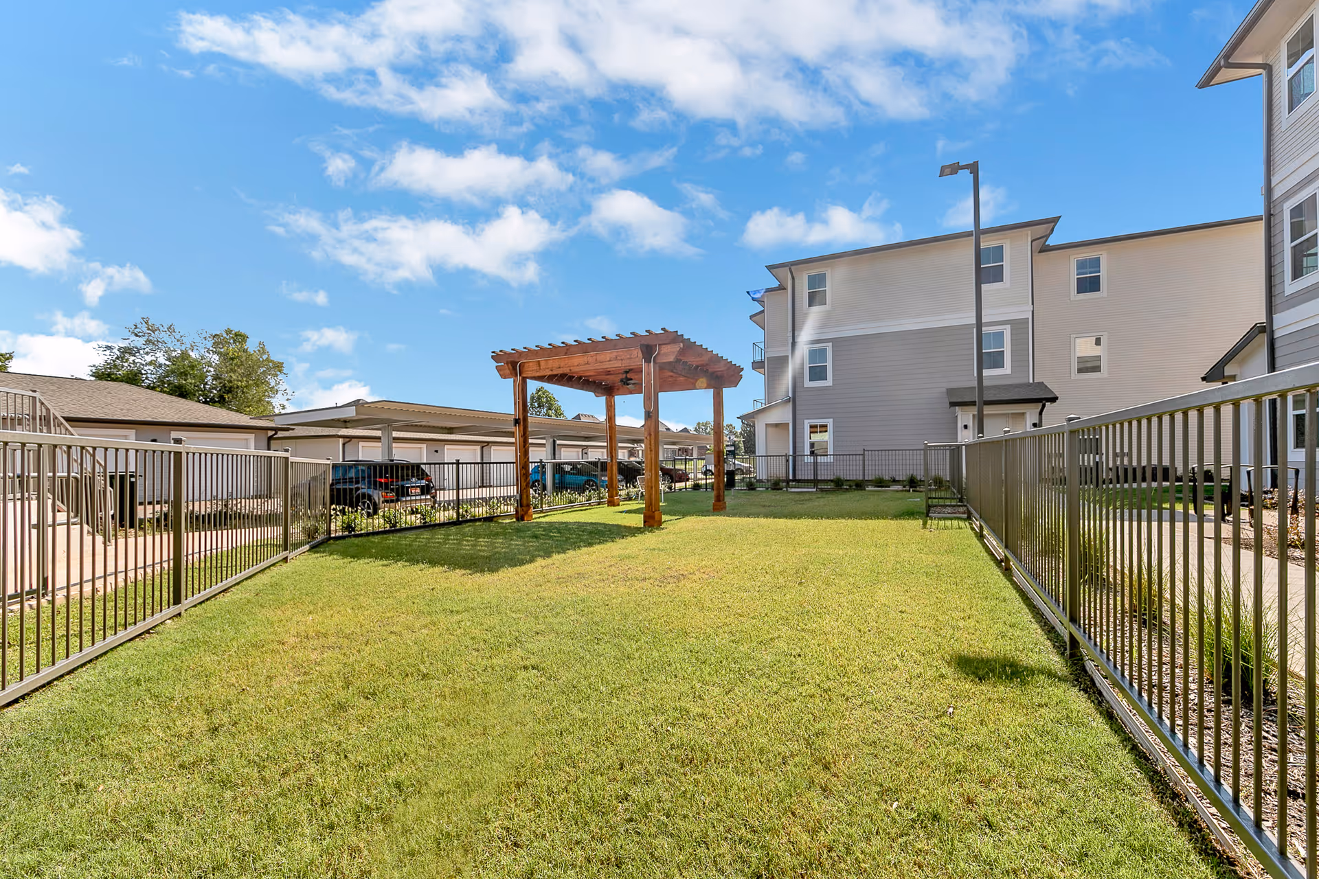 Outdoor grassy area enclosed by metal fencing with a wooden pergola in the center. Surrounding buildings include a multi-story residential building and a row of garages with parked cars. The sky is blue with scattered clouds.