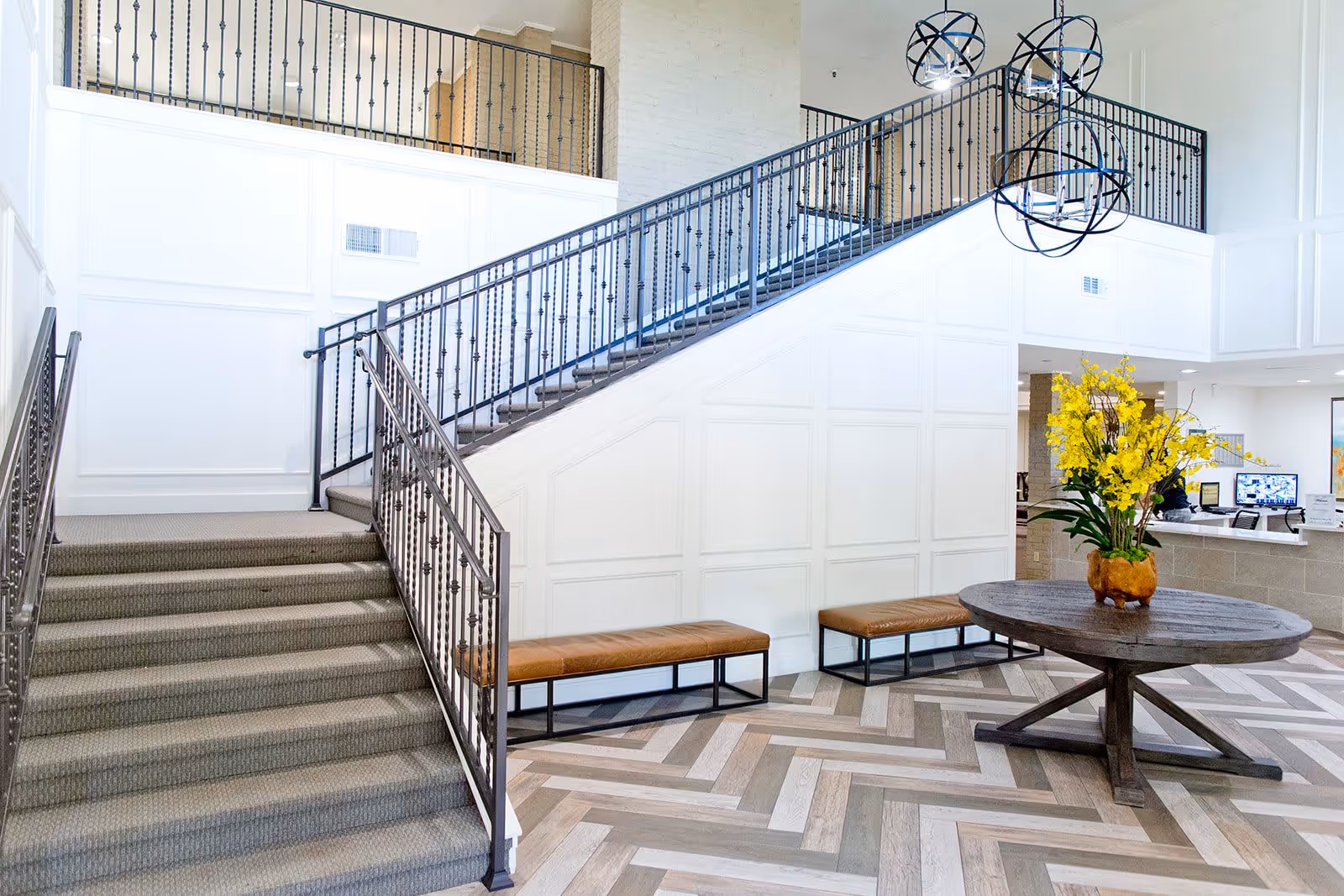 Bright lobby interior with a carpeted staircase and iron railing, a round wooden table with yellow flowers, and benches.
