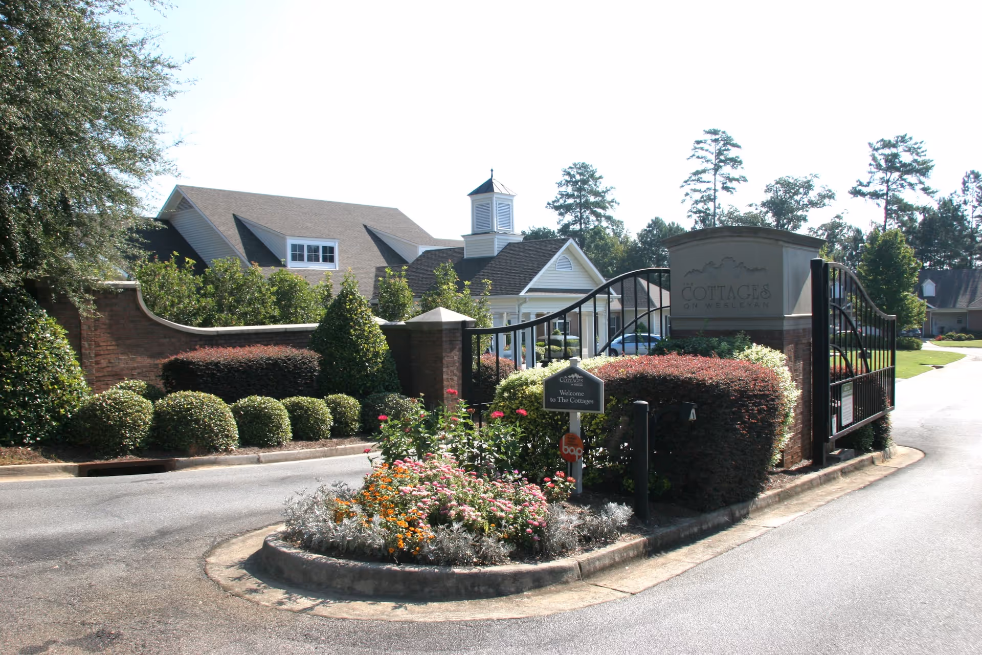 Entrance gate to The Cottages on Wesleyan senior living facility with a curved black metal gate, brick pillars, manicured bushes, and a flower bed in the center of the driveway. A sign on the right pillar reads 'Cottages on Wesleyan'.