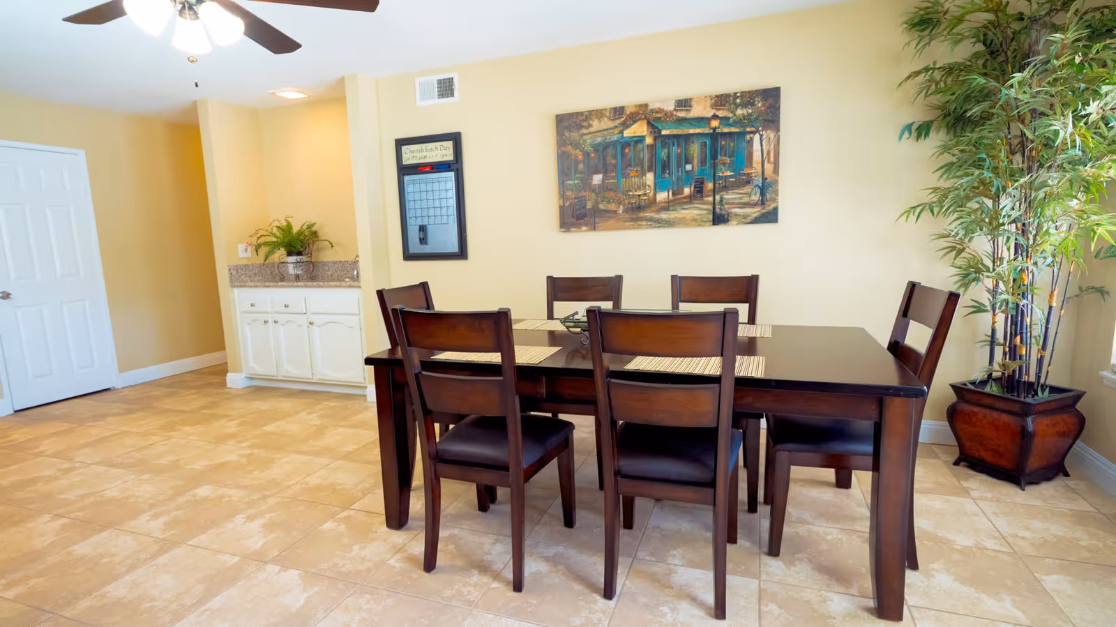 A dining area with a dark wooden table and six matching chairs on a tiled floor. The walls are painted light yellow, and there is a ceiling fan with lights above the table. On the wall, there is a framed calendar and a painting of a street scene with a café. A potted plant is placed in the corner near a window letting in natural light.
