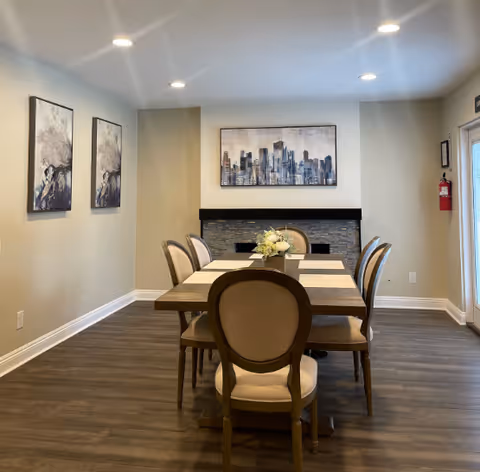 A dining room with a rectangular wooden table surrounded by six upholstered chairs. The table is set with placemats and a floral centerpiece. On the far wall, there is a modern fireplace with a stone surround and a cityscape painting above it. Two abstract paintings hang on the left wall. The room has recessed ceiling lights and wood flooring.