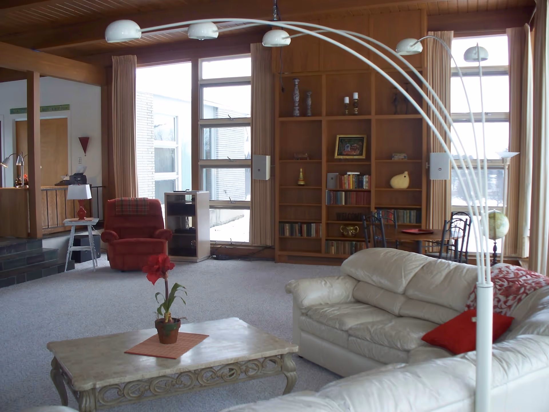 A spacious living room with a white leather sectional sofa adorned with red pillows and a red patterned throw. In front of the sofa is a decorative coffee table with a small potted plant. To the left, there is a red armchair next to a small white side table with a lamp. The room features large windows with beige curtains, a wooden bookshelf filled with books and decorative items, and a small round table with chairs near the windows. The ceiling and some walls are wooden, and the floor is carpeted in light gray.