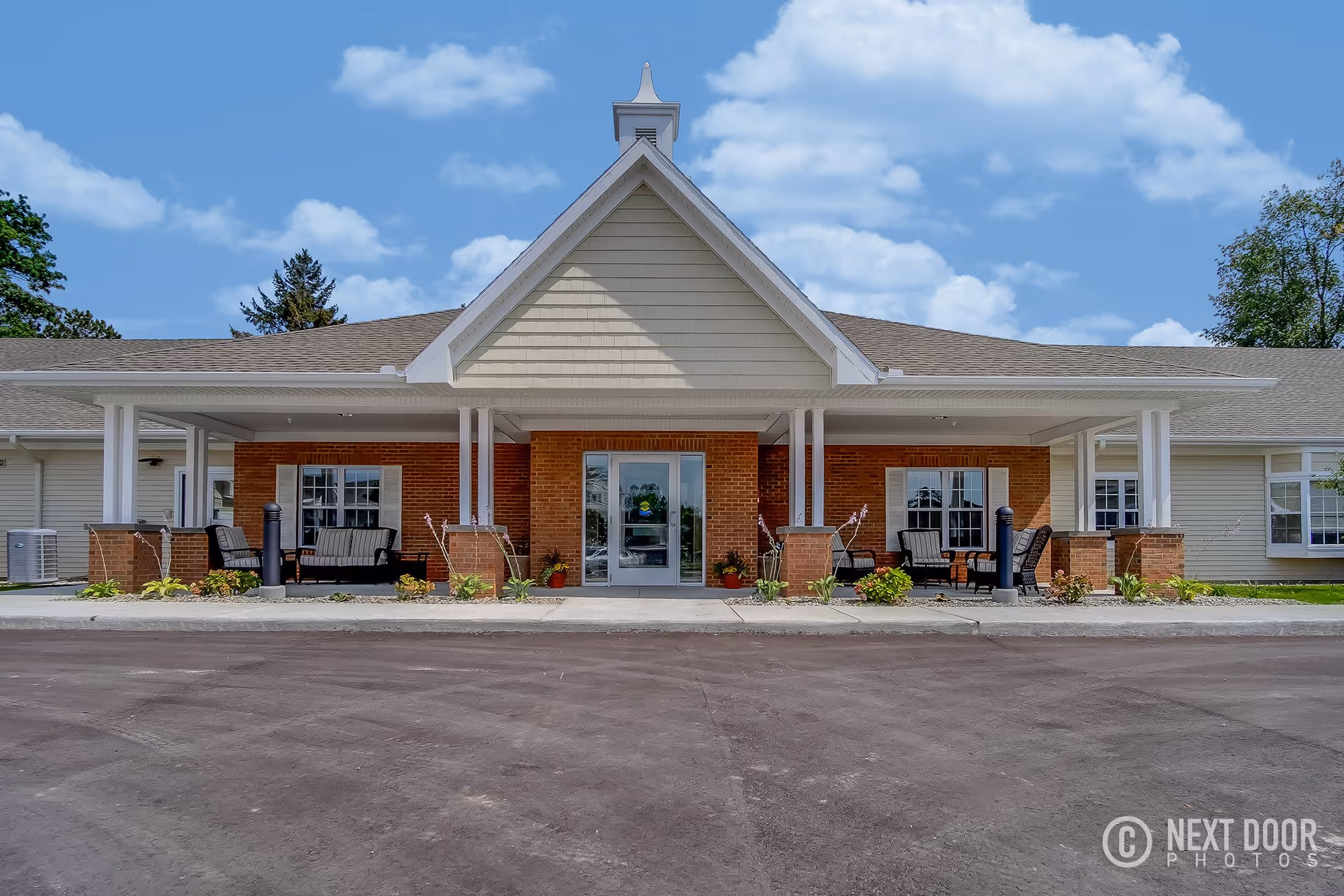 Front exterior view of Grandhaven Living Center showing a single-story building with a peaked roof, brick and siding facade, a covered entrance with seating areas on both sides, and a clear blue sky with some clouds.