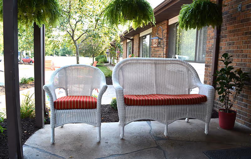 Outdoor seating area with a white wicker armchair and loveseat, both with red and white striped cushions, placed on a concrete patio in front of a brick building. There are hanging green plants above and a potted plant to the right, with trees and a parking lot visible in the background.