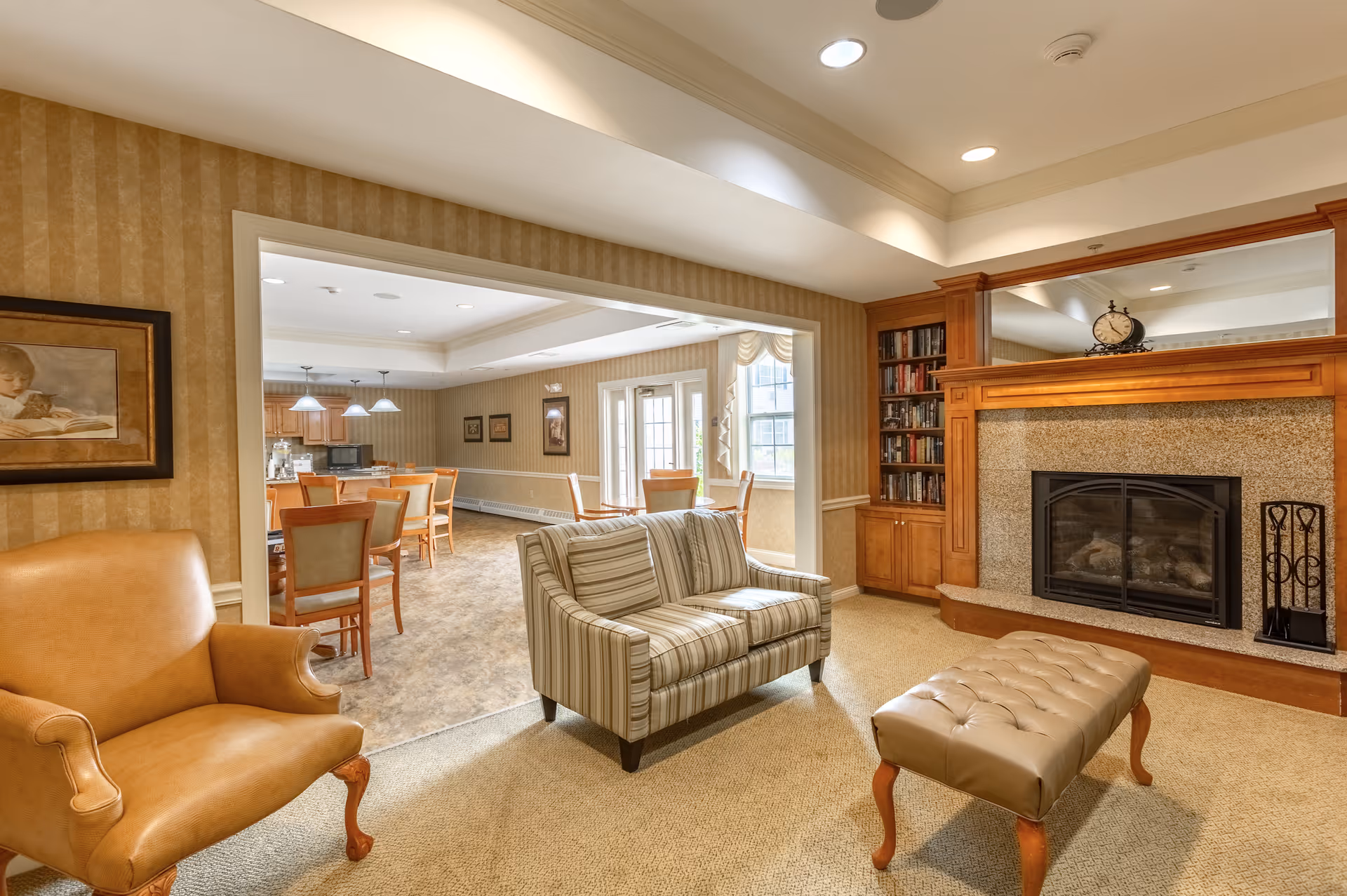 A cozy living room area with a striped loveseat, a tan leather armchair, and a tufted ottoman. The room features a fireplace with a wooden mantel and built-in bookshelves filled with books. The walls have a beige patterned wallpaper, and there is a large mirror above the fireplace. Through an open doorway, a dining area with wooden chairs and tables is visible, along with large windows letting in natural light.