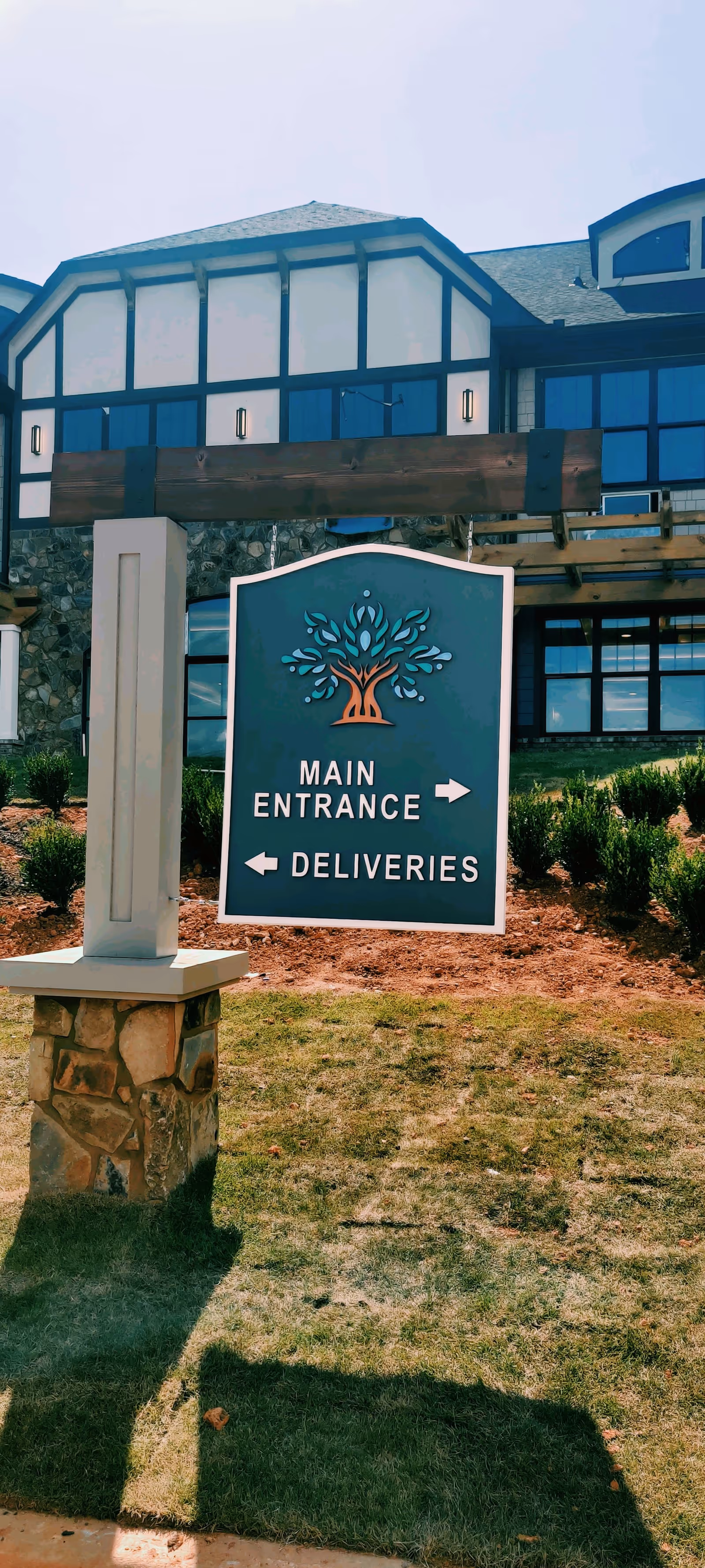 Outdoor view of a signpost in front of a building with a stone and wood facade. The sign features a tree logo and directional arrows indicating the main entrance to the right and deliveries to the left. The building has large windows and a landscaped area with grass and mulch.