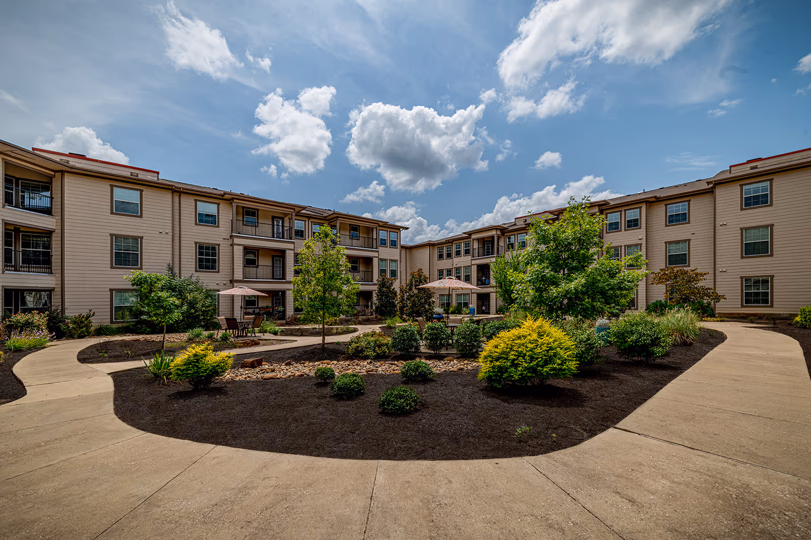 Outdoor courtyard area of a senior living facility with a curved concrete walkway surrounding landscaped garden beds with shrubs and small trees. The three-story beige building with multiple windows and balconies surrounds the courtyard under a partly cloudy blue sky.