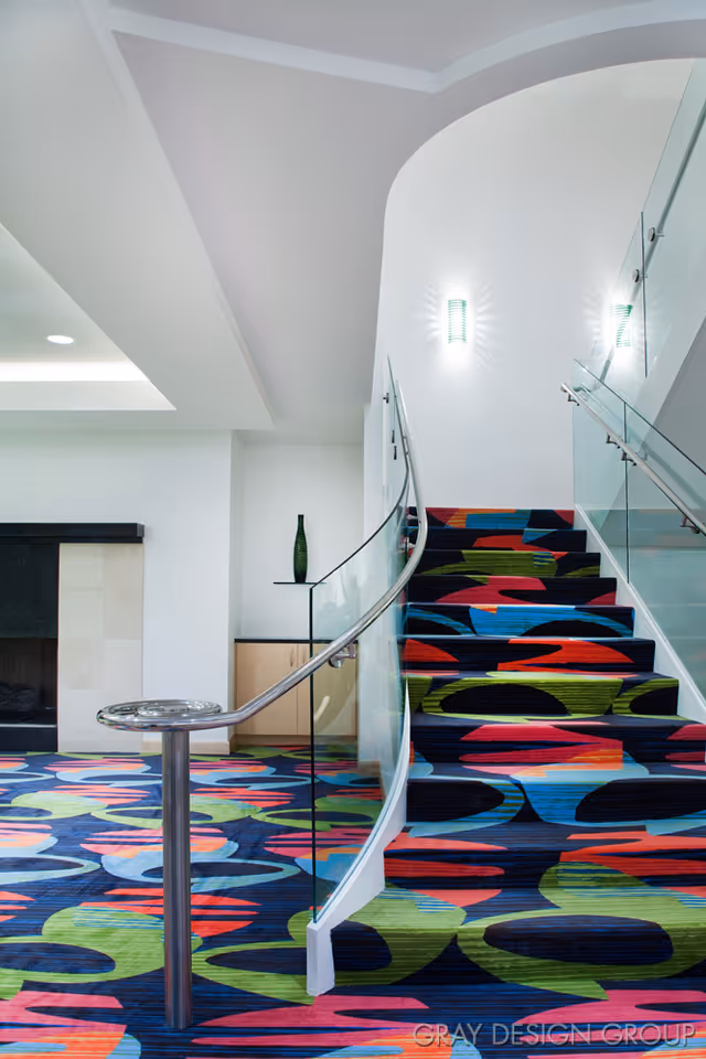 Interior view of a modern staircase with a colorful patterned carpet in shades of blue, green, red, and orange. The staircase has glass railings with metal handrails and leads up to a white wall with a wall-mounted light fixture. The floor below has the same colorful carpet pattern. There is a small cabinet with a green vase on top near the base of the stairs.