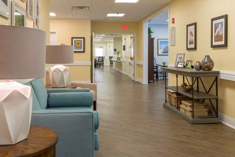 A well-lit hallway in a senior living facility with light yellow walls and wood flooring. On the left, there is a teal armchair with a side table and a lamp. On the right, a console table holds decorative items and framed photos, with framed artwork hanging above it. The hallway leads to a dining area with tables and chairs visible in the background.