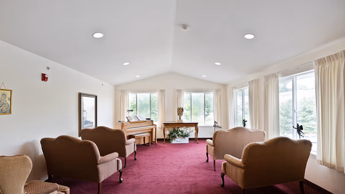 Bright communal sitting room with upholstered chairs, a piano, large windows and burgundy carpet.