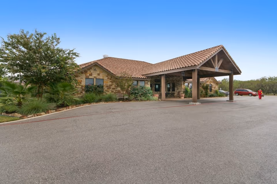 Exterior view of CelesteCare of Horseshoe Bay facility showing a single-story building with stone walls and a tiled roof. There is a covered entrance supported by wooden beams, surrounded by landscaped greenery including trees and shrubs. A red vehicle is parked in the driveway and a person dressed in red is walking nearby.