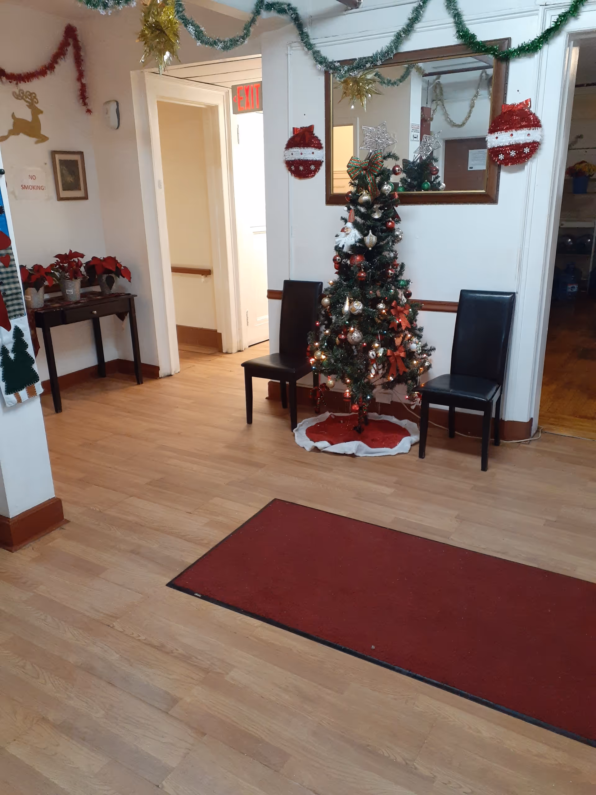 Interior hallway decorated for Christmas with a small decorated Christmas tree between two black chairs, garlands hanging from the ceiling, a red rug on the wooden floor, and a table with poinsettia plants. A mirror is mounted on the wall above the tree, reflecting part of the decorations.
