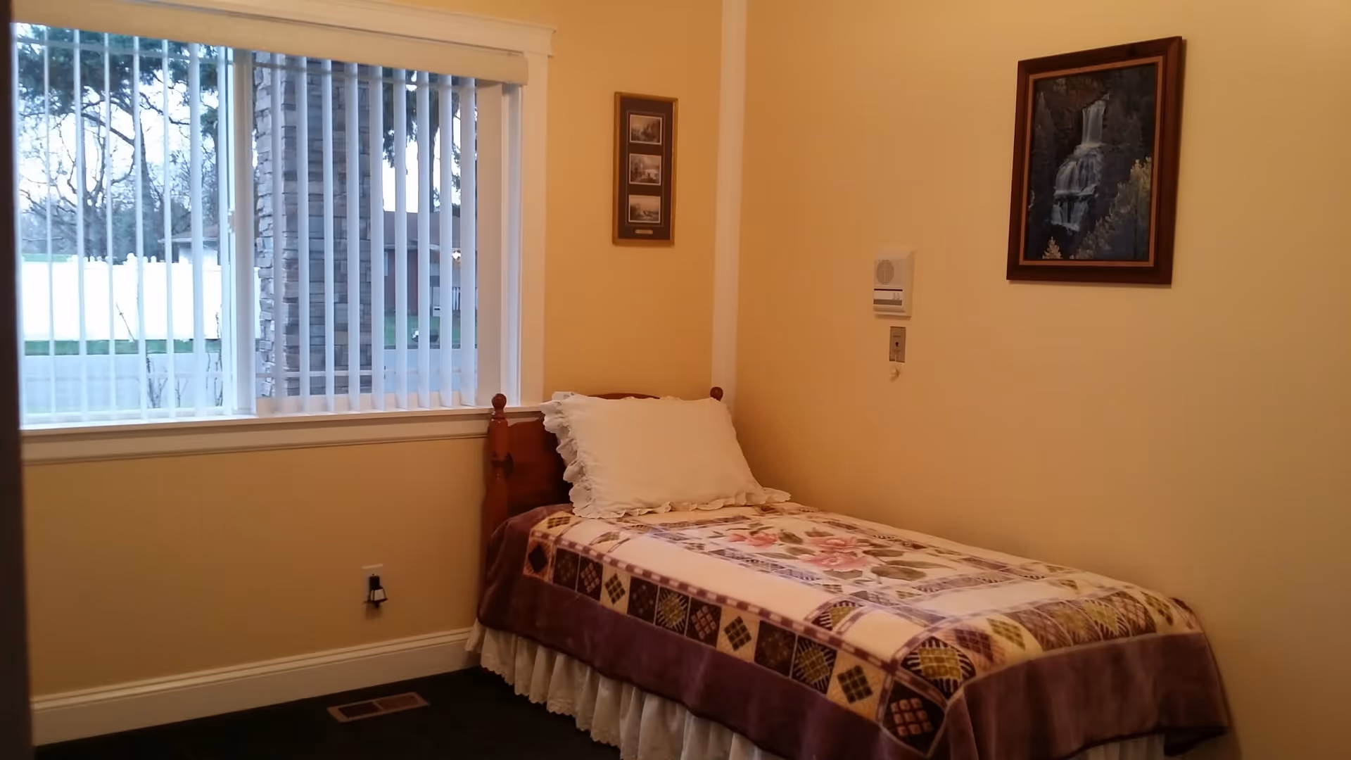 A small bedroom with a single wooden bed against a beige wall. The bed has a floral and patchwork quilt with a white pillow. There is a window with vertical blinds on the left side, showing an outdoor view with a white fence and trees. Two framed pictures hang on the walls, one above the bed and one to the left of it.