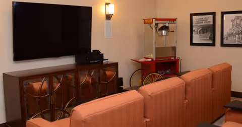 A cozy room with a large flat-screen TV mounted on the wall above a decorative wooden cabinet. In front of the TV, there are several orange cushioned chairs arranged in rows. To the right, there is a red popcorn machine on a stand with yellow wheels. The walls are decorated with framed black and white pictures and a wall-mounted light fixture.