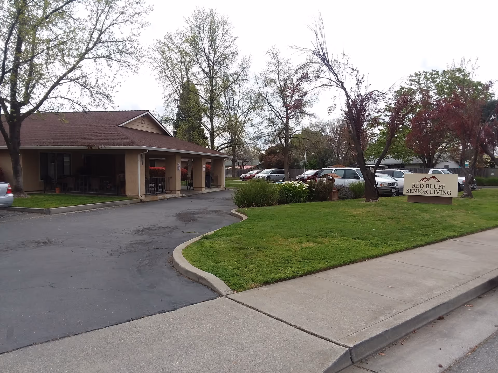 Exterior view of Red Bluff Senior Living facility showing a single-story building with a covered porch, a parking area with several cars, green lawn, trees, and a sign that reads 'Red Bluff Senior Living'.