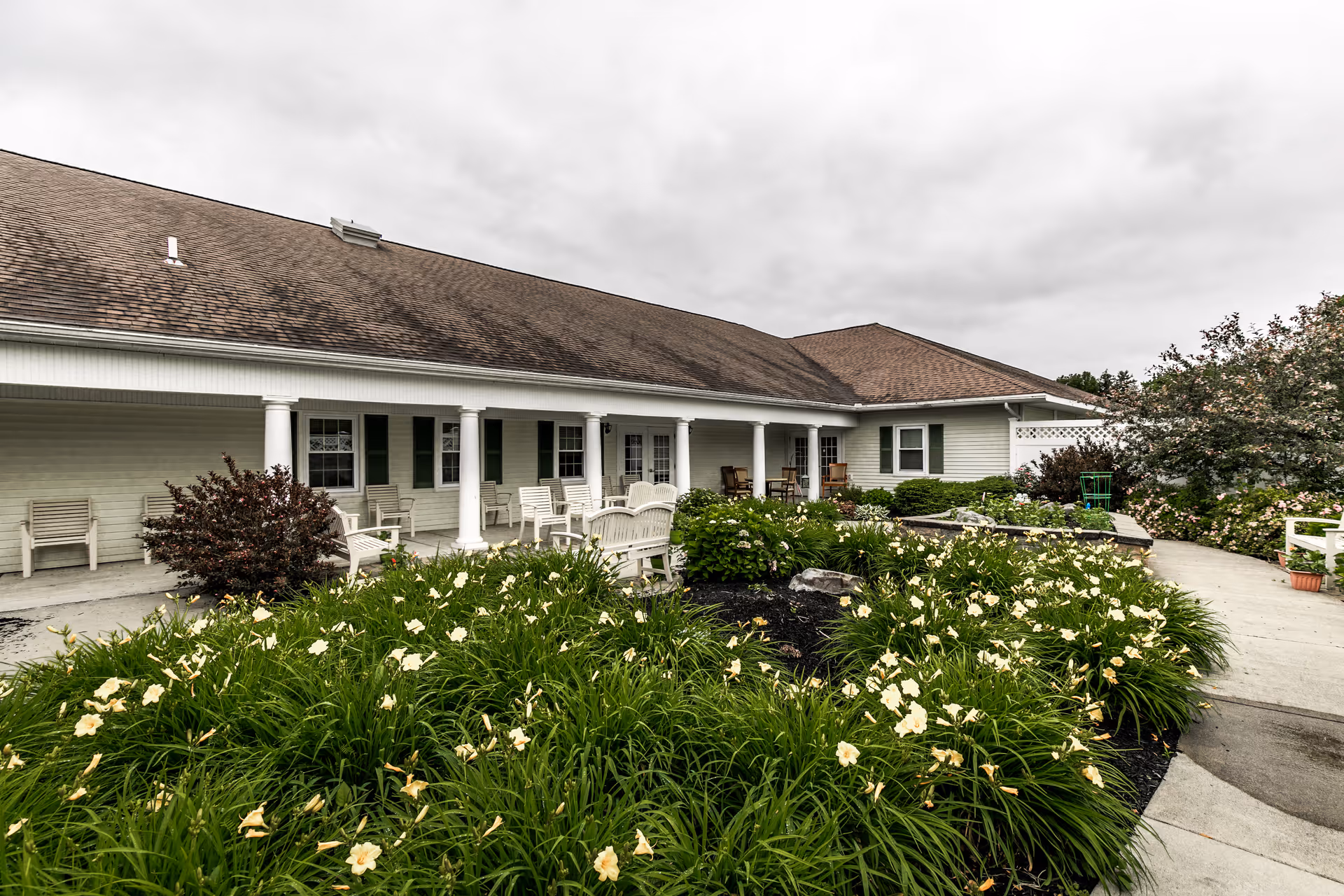 Front exterior of a single-story senior living building with a covered porch, chairs, and landscaped flower beds.