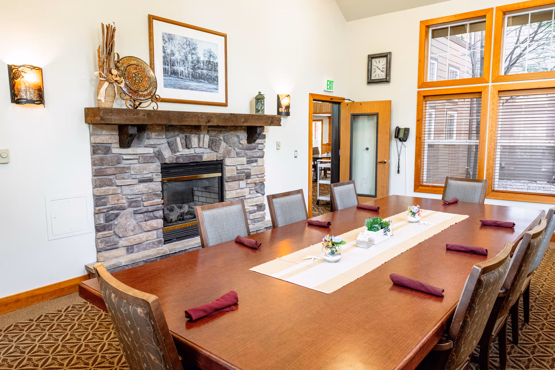 A dining room with a long wooden table set with burgundy napkins and small decorative plants. The room features a stone fireplace with a wooden mantel, a framed black and white photograph above it, and large windows with wooden frames letting in natural light. There is a clock on the wall and a door leading to another room.