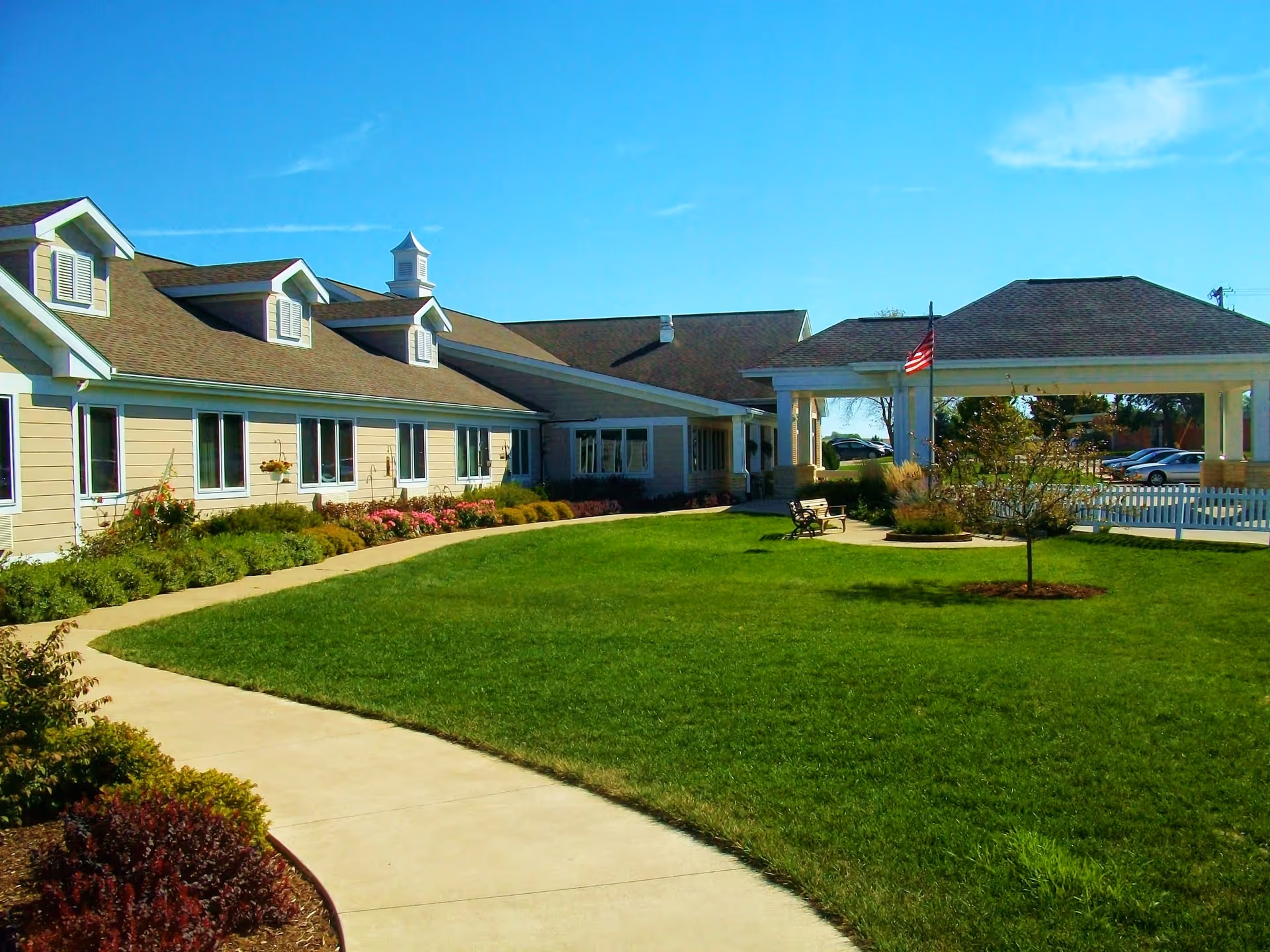 Front exterior of a single-story senior living facility with a green lawn, curved walkway, covered entrance, benches, and an American flag.