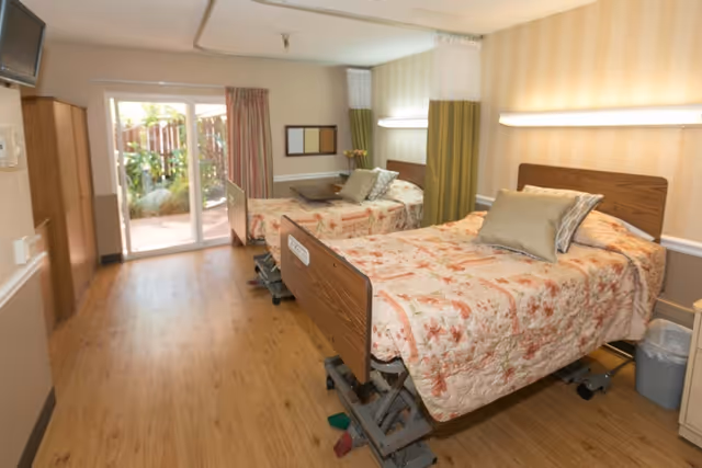 Two hospital-style beds with floral bedspreads in a bright nursing facility room with wood flooring and a sliding glass door to an outdoor patio.