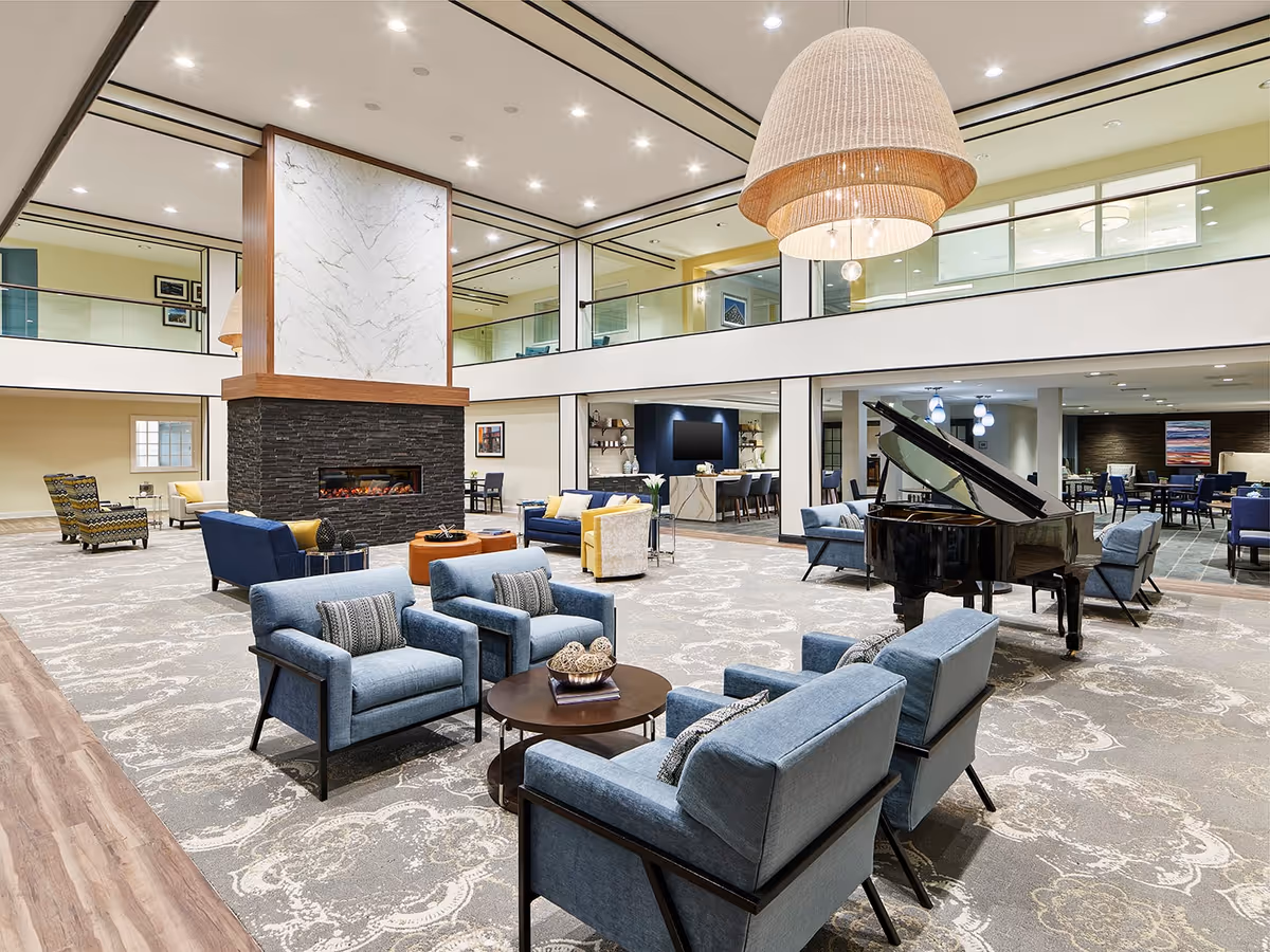 Spacious and modern senior living facility common area with a large two-sided fireplace in the center, surrounded by comfortable seating including blue armchairs and sofas. A grand piano is positioned to the right, and there is a dining area visible in the background. The space features high ceilings with recessed lighting and a second-floor balcony overlooking the area.