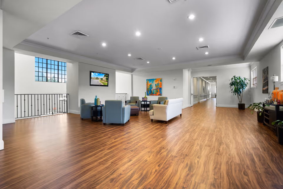 A spacious and well-lit common area with wood flooring, several armchairs and a small sofa arranged around a coffee table. The walls are white with colorful artwork and a flat-screen TV mounted on one wall. There are large windows letting in natural light, and a hallway leading to other rooms. Potted plants and a sideboard with decorative items are also visible.
