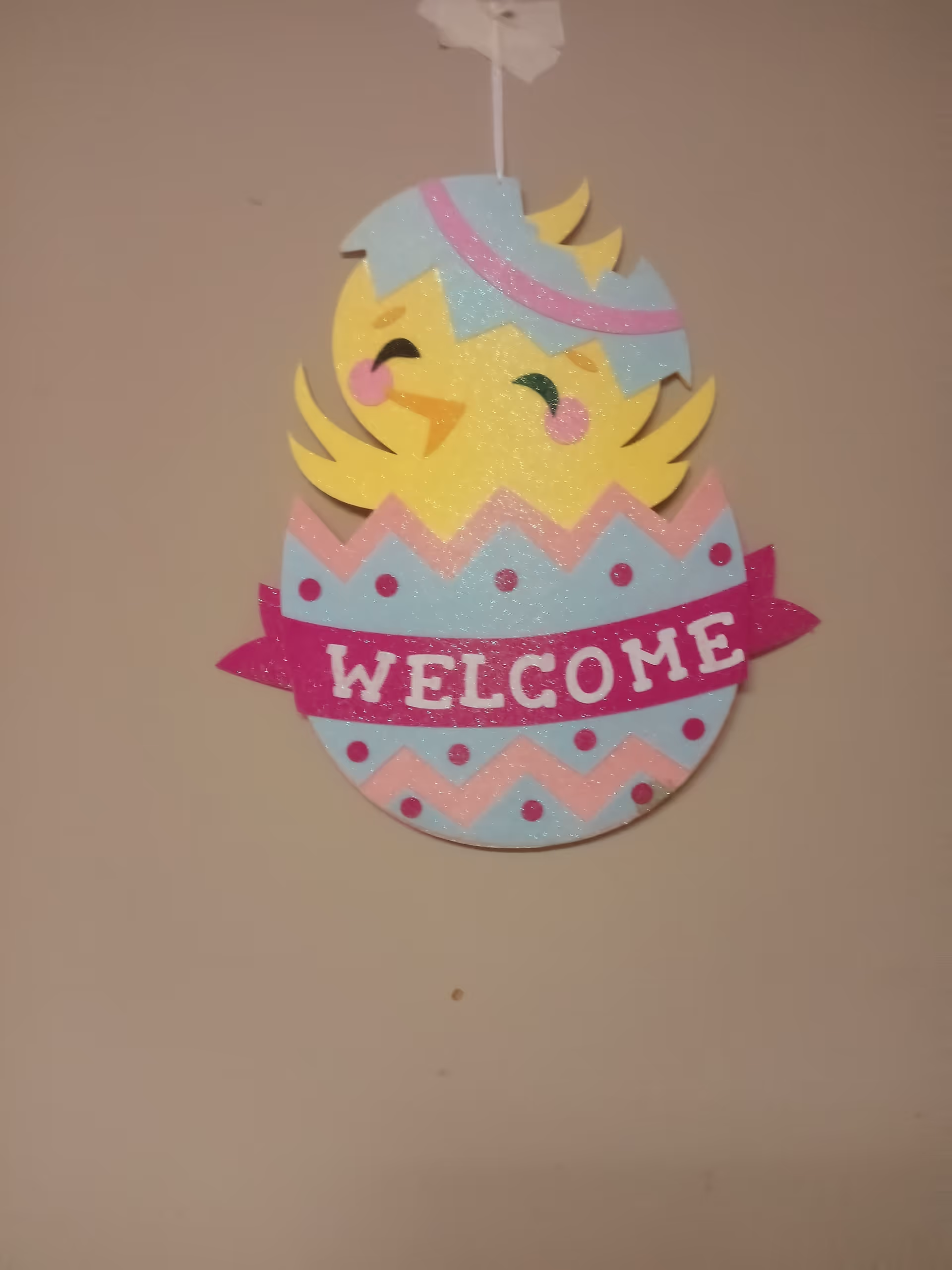 Colorful glittery hanging welcome sign shaped like a chick hatching from an egg on a beige wall.
