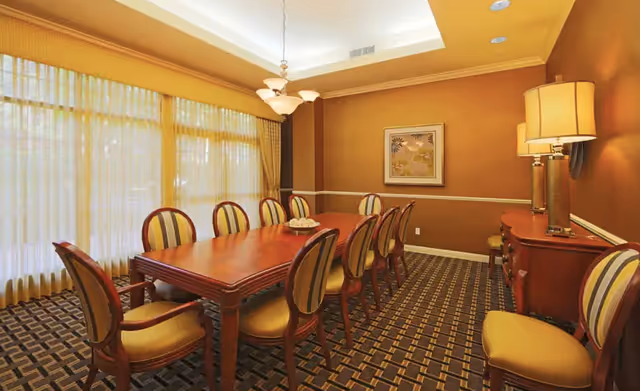 A formal dining room with a long wooden table surrounded by eight upholstered chairs with striped fabric. The room has warm brown walls, a patterned carpet, a chandelier hanging from the ceiling, a sideboard with a lamp, and a framed floral artwork on the wall. Large windows with sheer curtains allow natural light to fill the space.