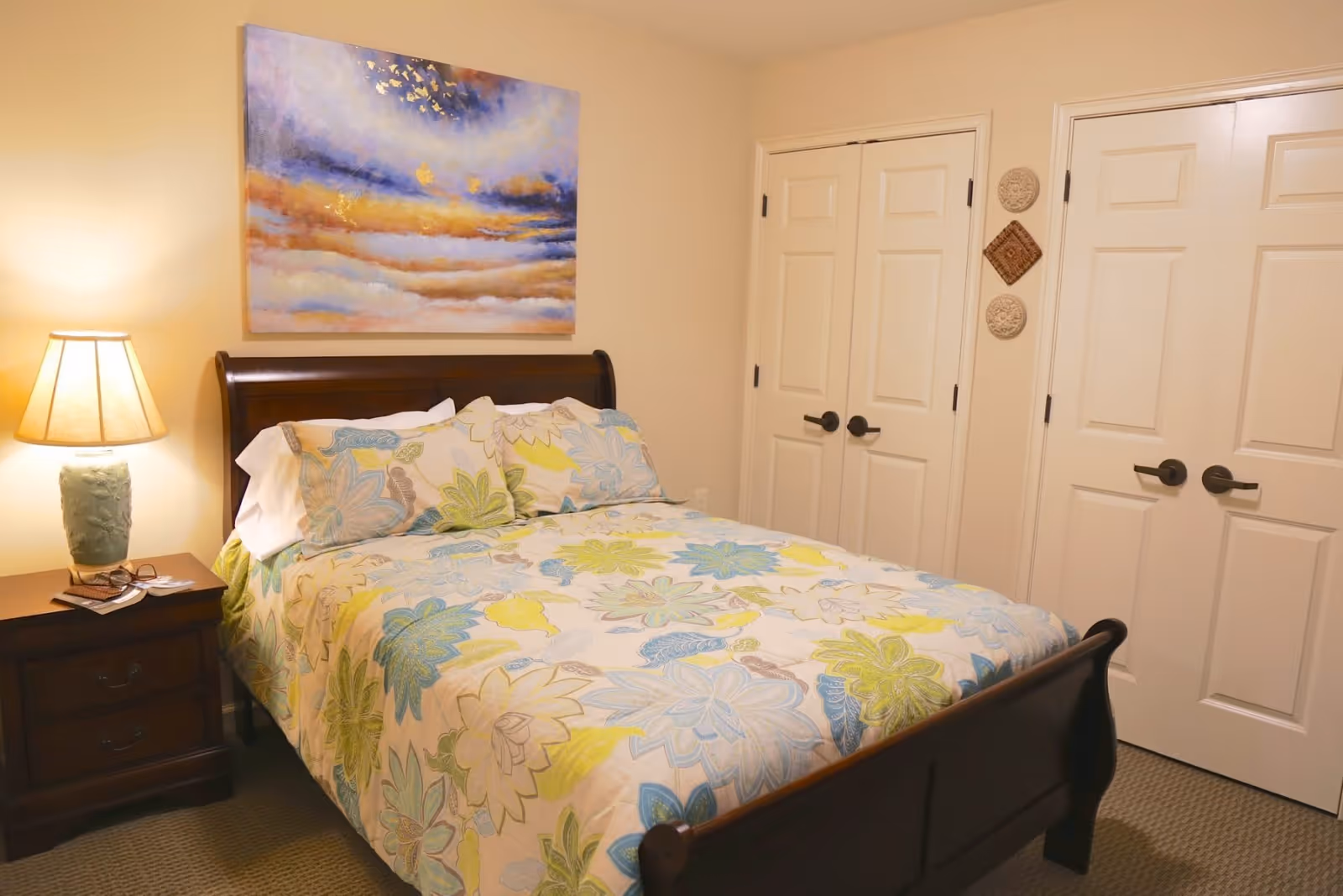 A cozy bedroom with a wooden bed frame and a floral patterned bedspread in shades of blue, green, and yellow. There is a wooden nightstand with a lamp and some reading materials on it. Above the bed hangs a colorful abstract painting. The room has beige walls and carpeted floor, with two closed white doors on the right side.