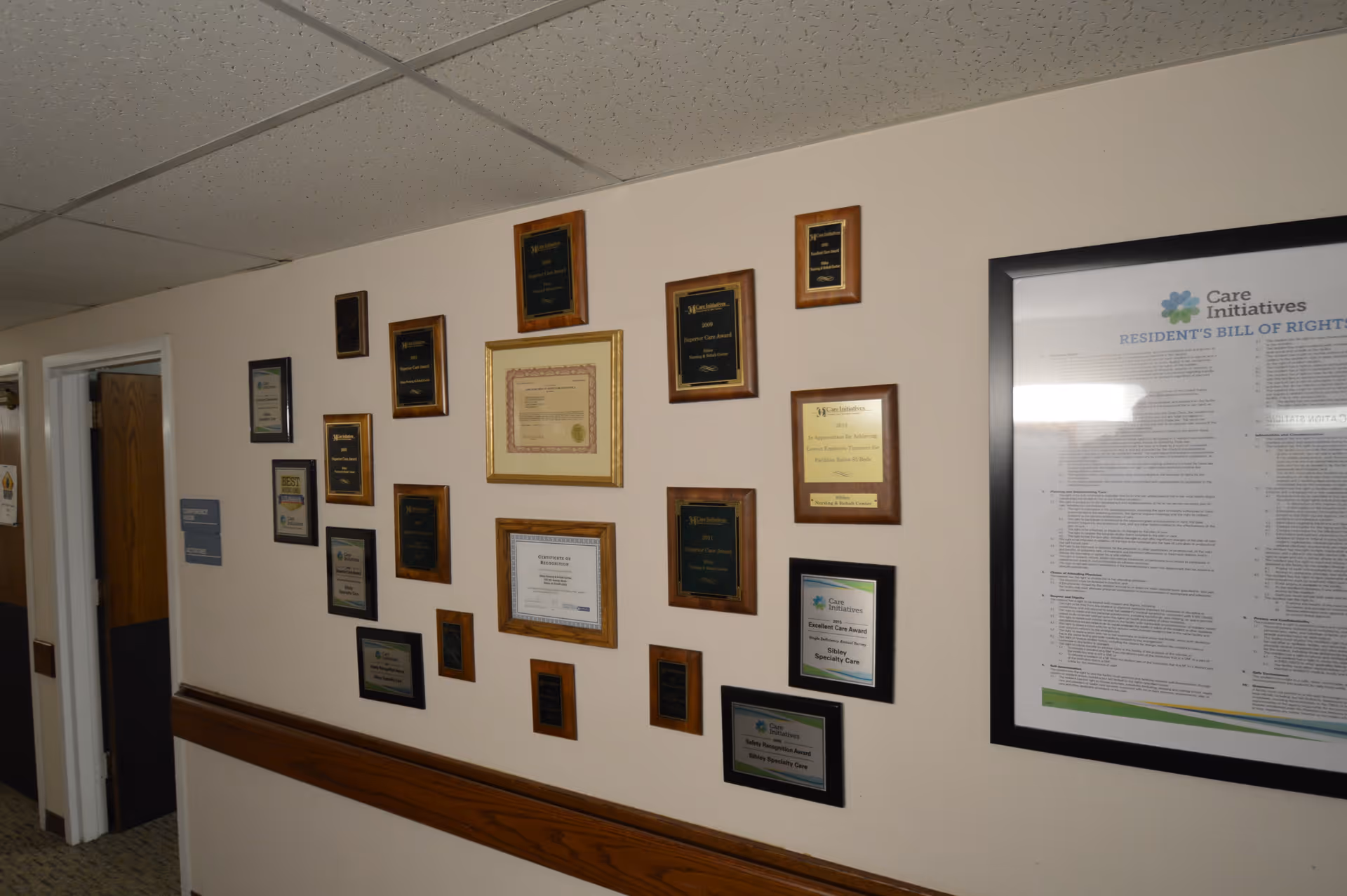 A hallway wall decorated with multiple framed awards and certificates, including plaques and a large framed document titled 'Resident's Bill of Rights' from Care Initiatives. The wall has a wooden chair rail and a drop ceiling with fluorescent lighting.
