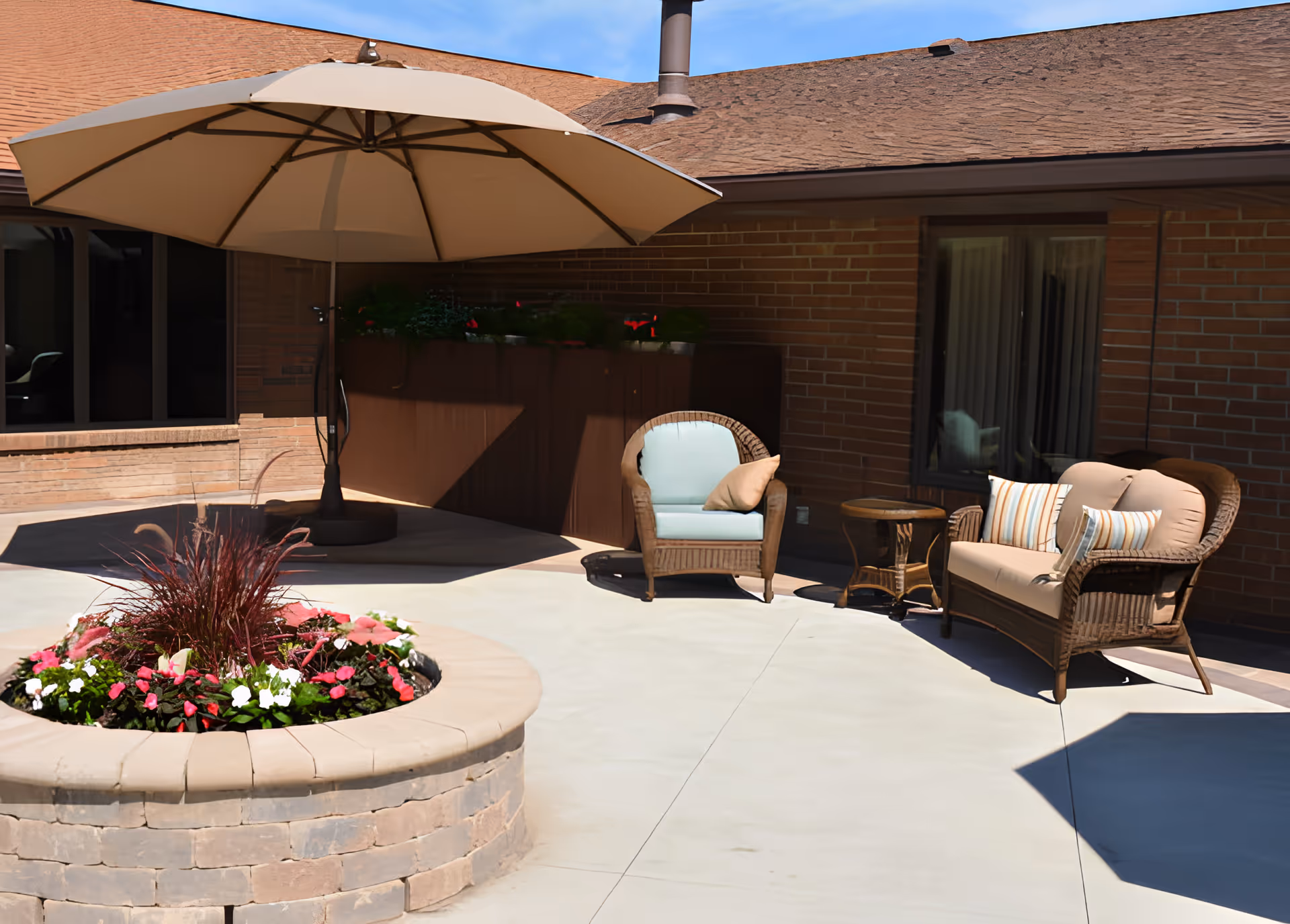 Outdoor patio area with a large beige umbrella, wicker chairs with cushions, a small wicker table, and a circular raised flower bed with colorful flowers and plants. The patio is adjacent to a brick building with windows.