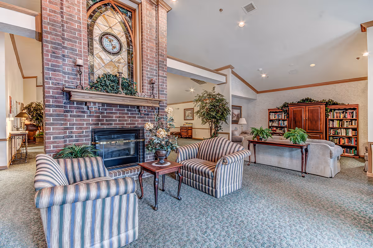 A cozy living room area with a brick fireplace featuring a decorative stained glass window above it. Two striped armchairs and a small wooden table with a floral arrangement are positioned in front of the fireplace. In the background, there is a beige sofa, a wooden console table with plants, bookshelves filled with books, and framed artwork on the walls. The room has carpeted floors and soft lighting.