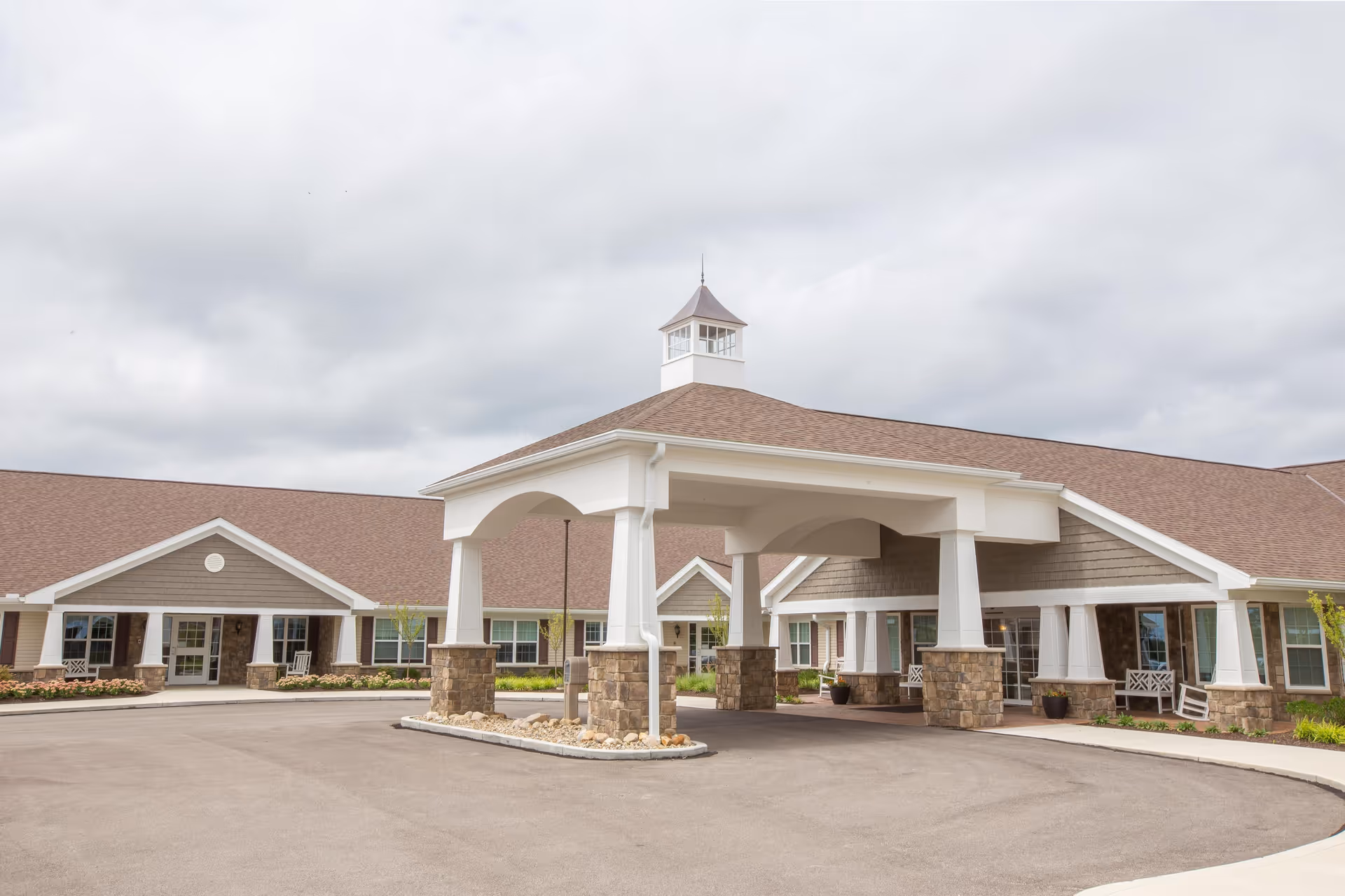 Exterior view of a single-story senior living facility with a large covered entrance supported by white columns with stone bases, a circular driveway, and a cloudy sky overhead.