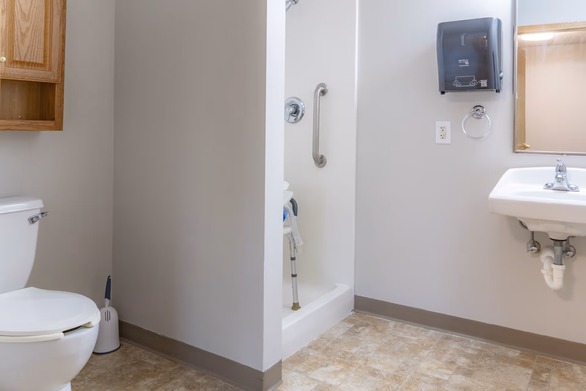 A clean bathroom with a white toilet, a small shower with a grab bar and a shower chair, a white sink with a mirror above it, a paper towel dispenser, and a wooden cabinet mounted on the wall.