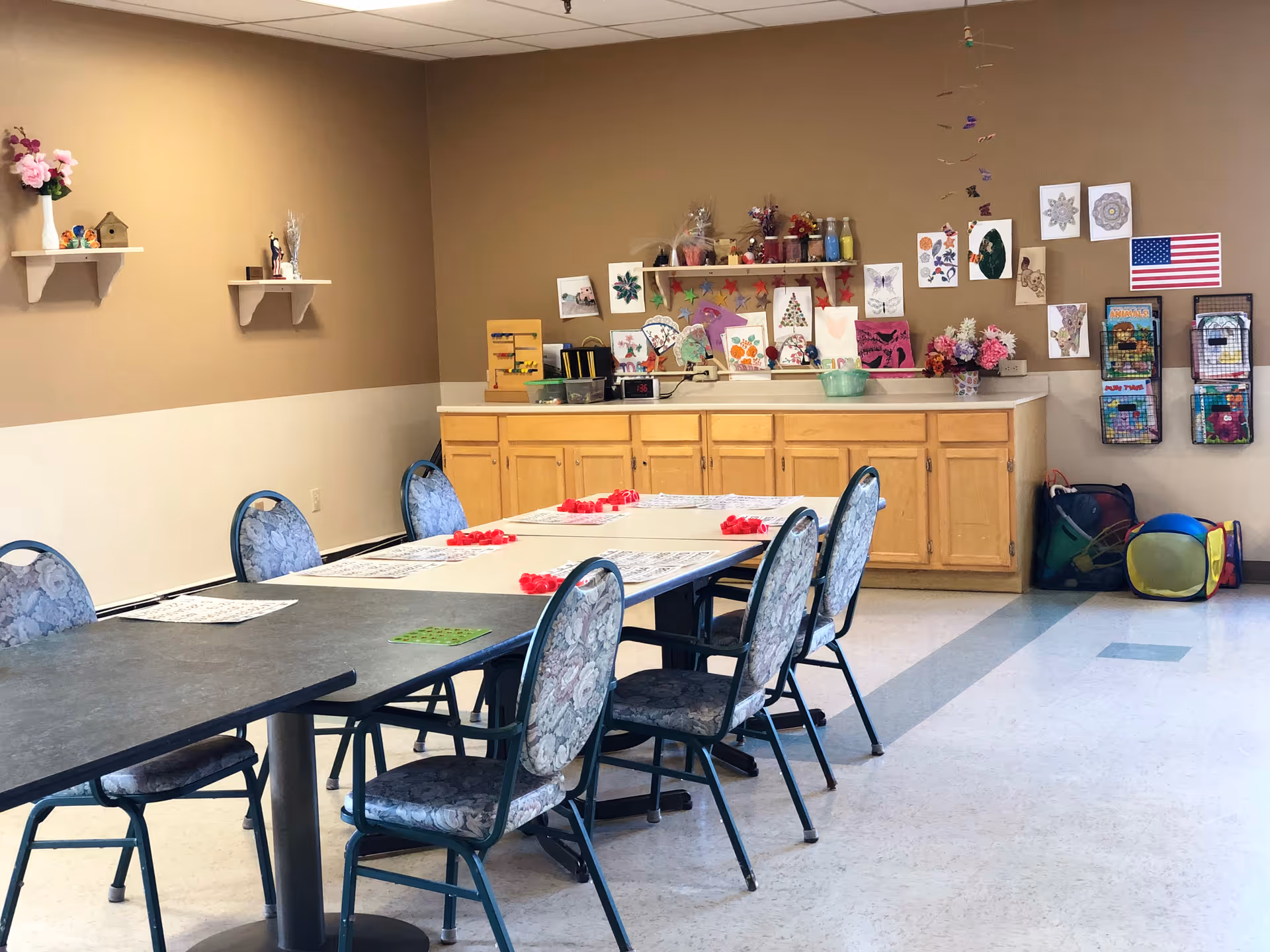A well-lit activity room with a long table surrounded by six floral-patterned chairs. The table has placemats and red game pieces arranged on it. The back wall features wooden cabinets topped with various craft supplies, artwork, and decorations. Shelves on the left wall hold small decorative items, and the right wall has an American flag, wall-mounted magazine holders, and some colorful play items on the floor.