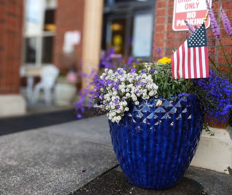 Blue ceramic planter filled with white and purple flowers and a small American flag at a brick building entrance.