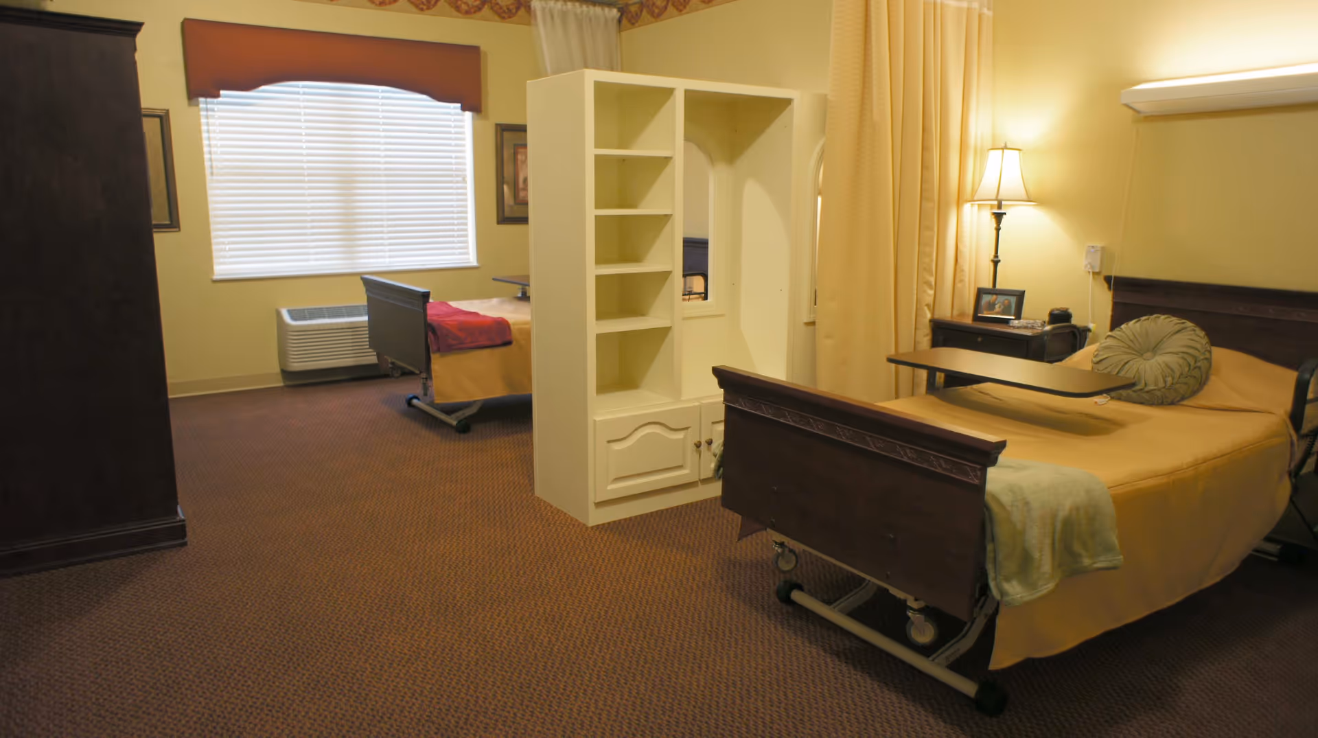 A spacious senior bedroom with two hospital-style beds separated by a white shelving divider, a nightstand and lamp.