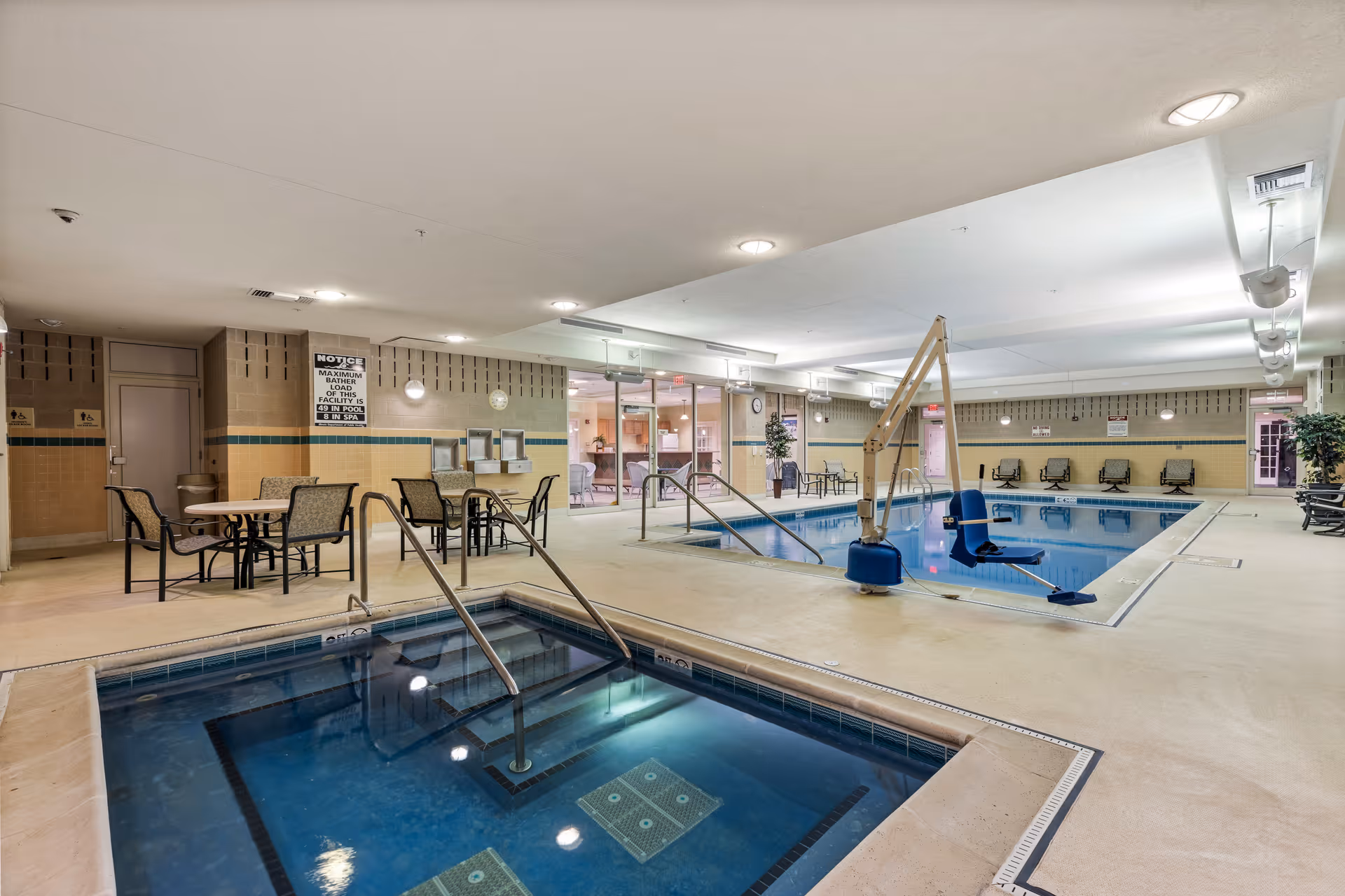 Indoor pool area with a swimming pool and a smaller hot tub in the foreground. There are several chairs and tables around the pool, and a pool lift chair for accessibility. The walls are tiled with beige and green accents, and there are glass doors leading to another room in the background.