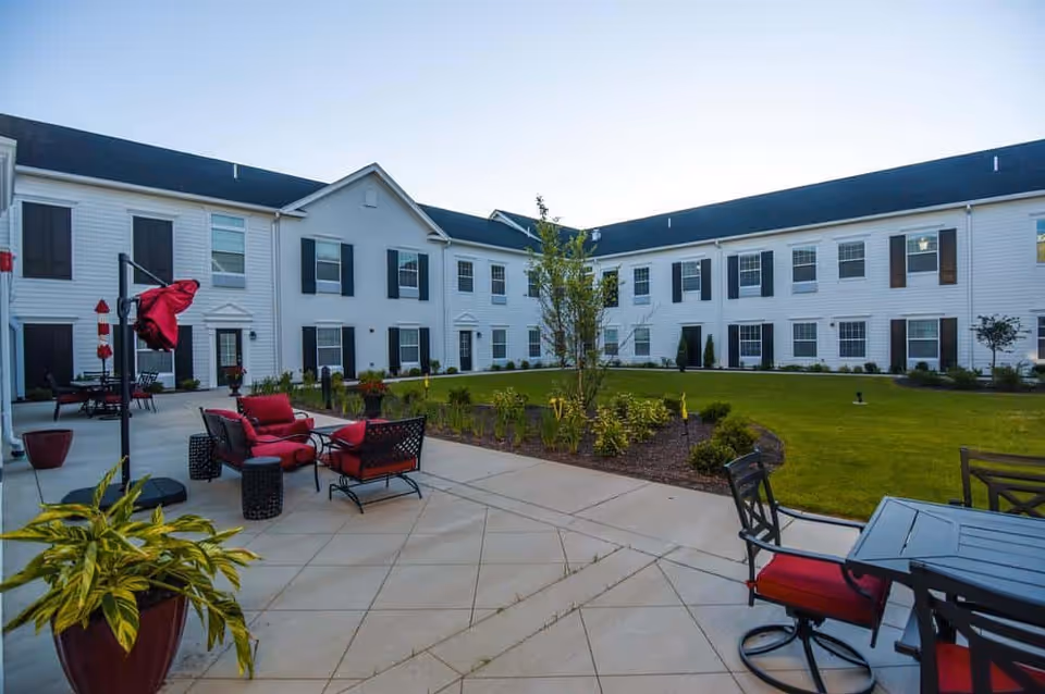 Landscaped courtyard with patio seating and red-cushioned chairs surrounded by a two-story white senior living building.