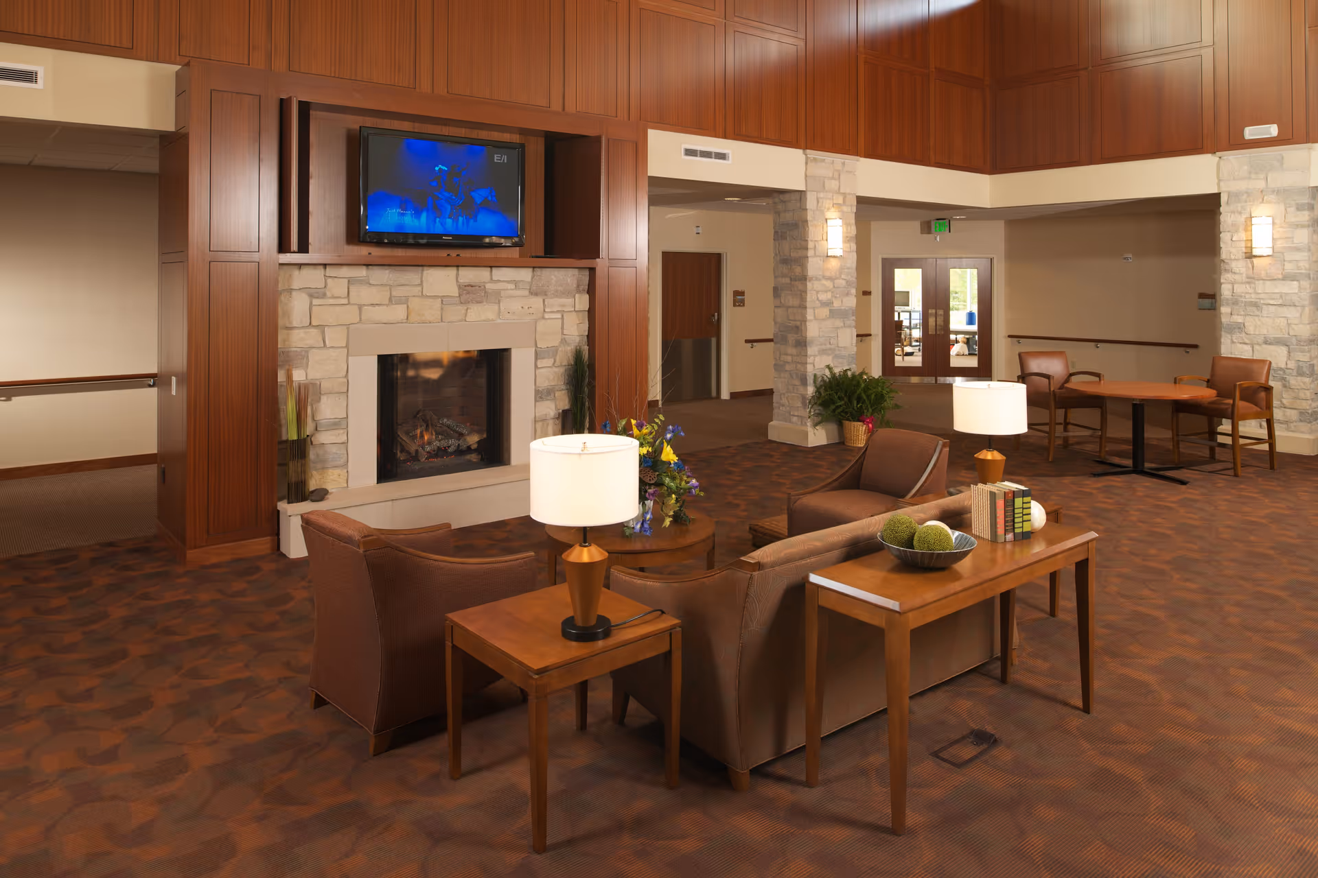 A cozy living room area in a senior living facility featuring a stone fireplace with a mounted flat-screen TV above it. The room has brown upholstered chairs and a sofa arranged around a wooden coffee table with a lamp and decorative items. The walls are paneled with wood and stone columns, and there are additional seating areas with tables and chairs in the background. The space is warmly lit and carpeted with a patterned design.
