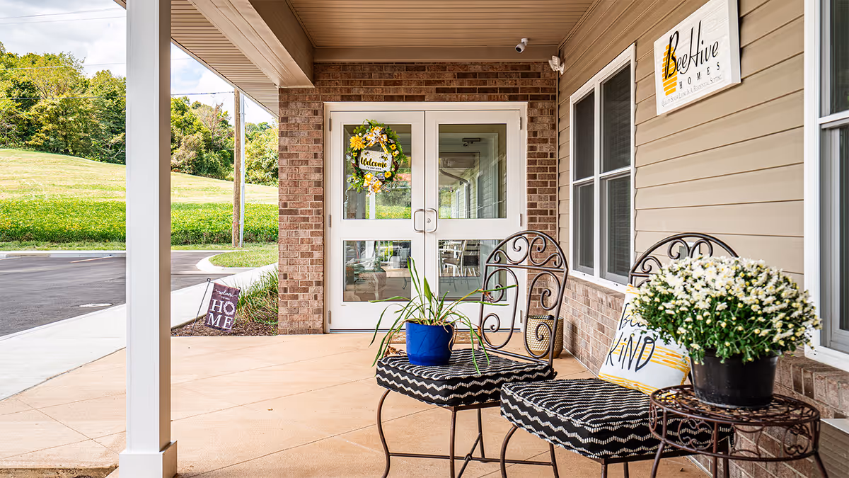Front porch area of BeeHive Homes of Taylorsville featuring two black wrought iron chairs with patterned cushions, a small round table with a potted white flower, a blue pot with a green plant on one chair, a welcome wreath on the glass double doors, and a sign on the wall with the BeeHive Homes logo.