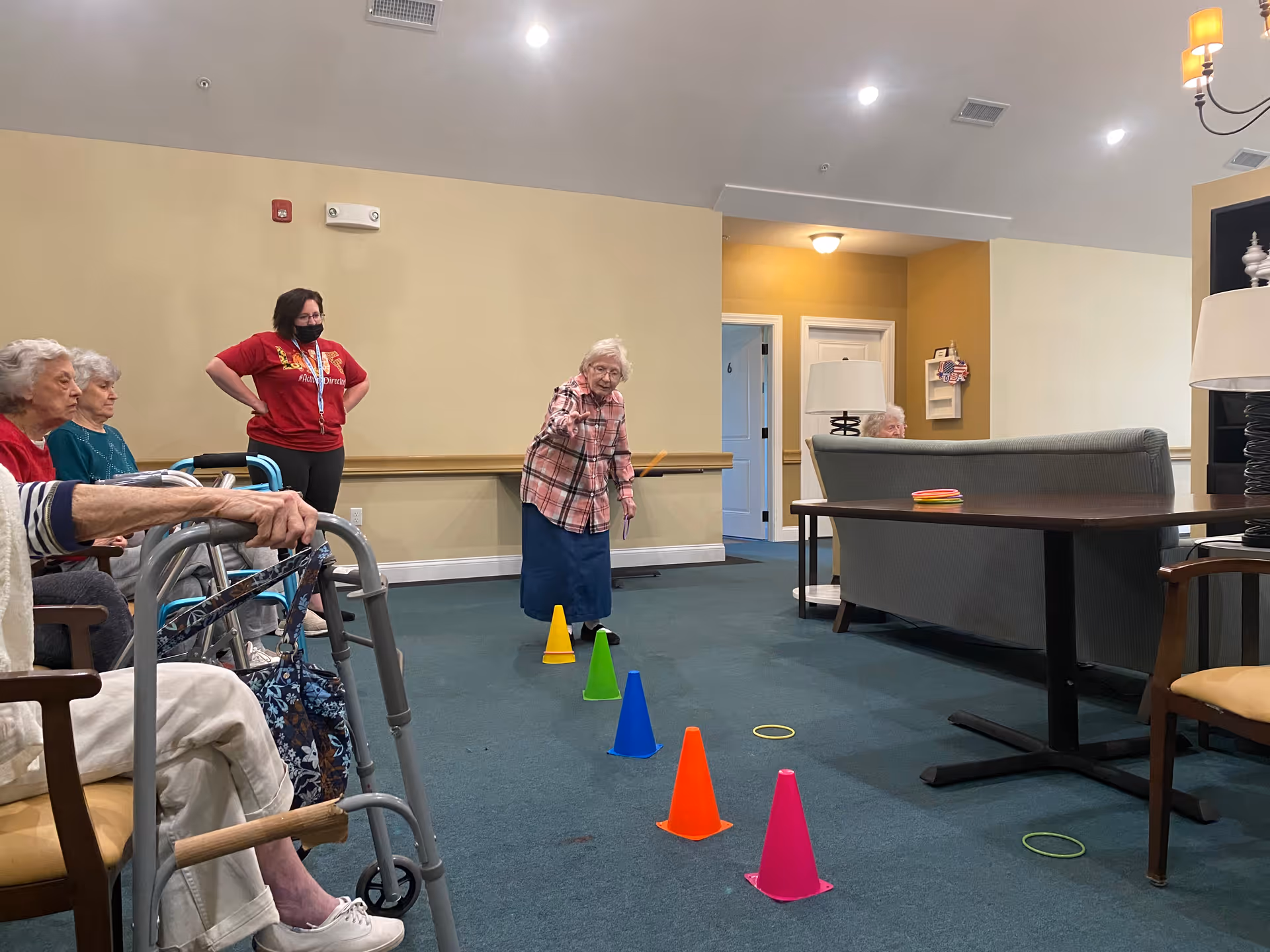 A group of elderly women and a caregiver in a red shirt and black mask are in a common area with carpeted floor and beige walls. One elderly woman is standing and appears to be tossing a ring towards colorful cones arranged in a line on the floor. Other elderly women are seated, some with walkers, watching the activity. The room has chairs, tables, and lamps, creating a cozy atmosphere.