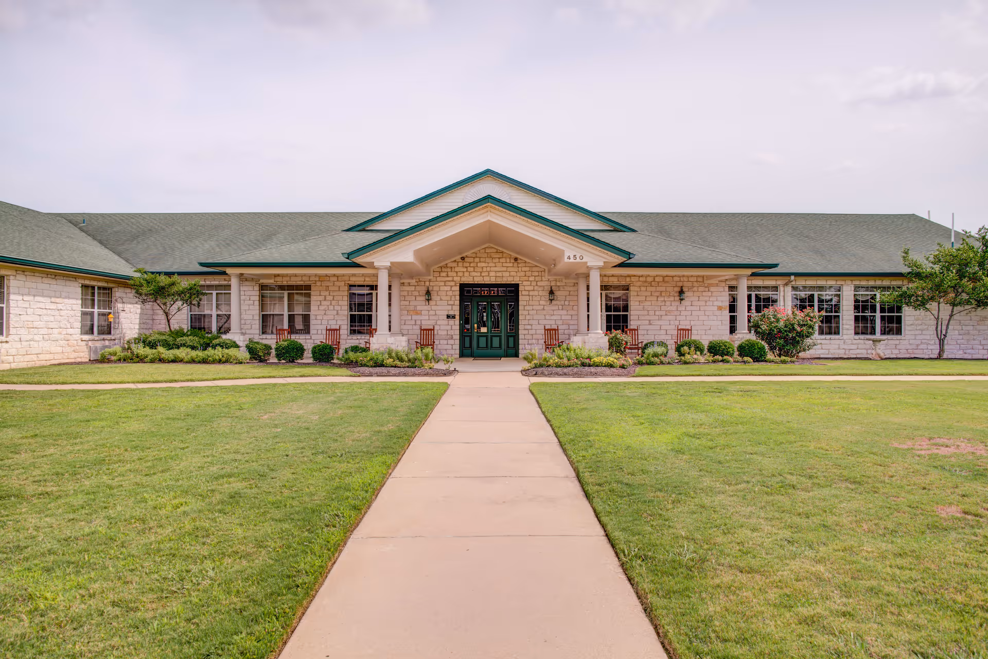 Front exterior view of The Pointe at Cedar Park building with a green roof, stone facade, a central entrance with columns, and a walkway leading up to the door. The surrounding area has well-maintained grass and shrubs.