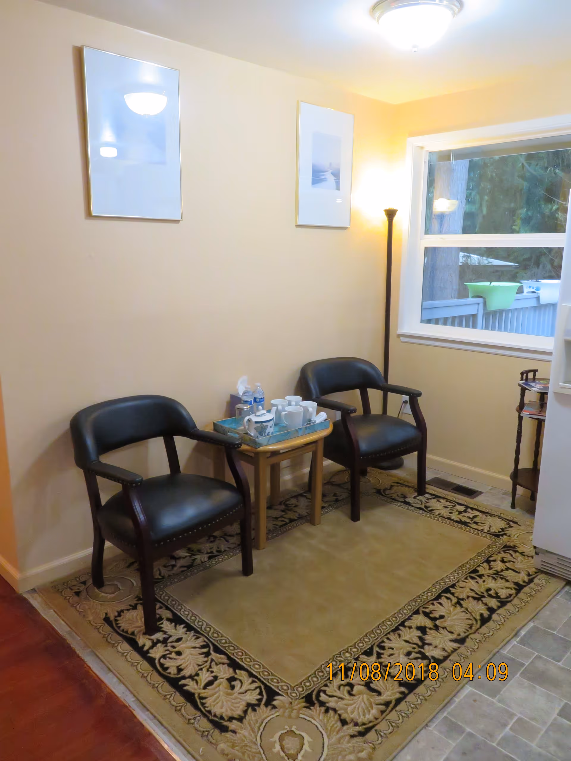 A small seating area with two black leather armchairs separated by a wooden side table holding a tray with cups, a teapot, and water bottles. The area is carpeted with a decorative rug and has beige walls with two framed pictures. A window shows an outdoor view with trees and a green bowl on a railing. A floor lamp is in the corner providing light.