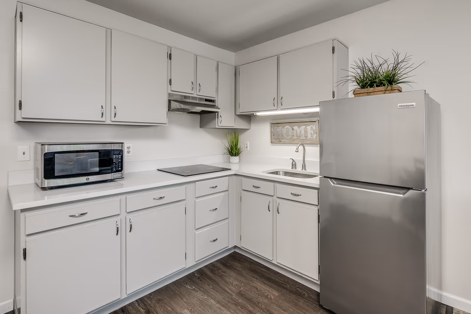 Compact bright kitchen with white cabinets, a stainless steel refrigerator, microwave, sink, and a decorative "HOME" sign above the counter.